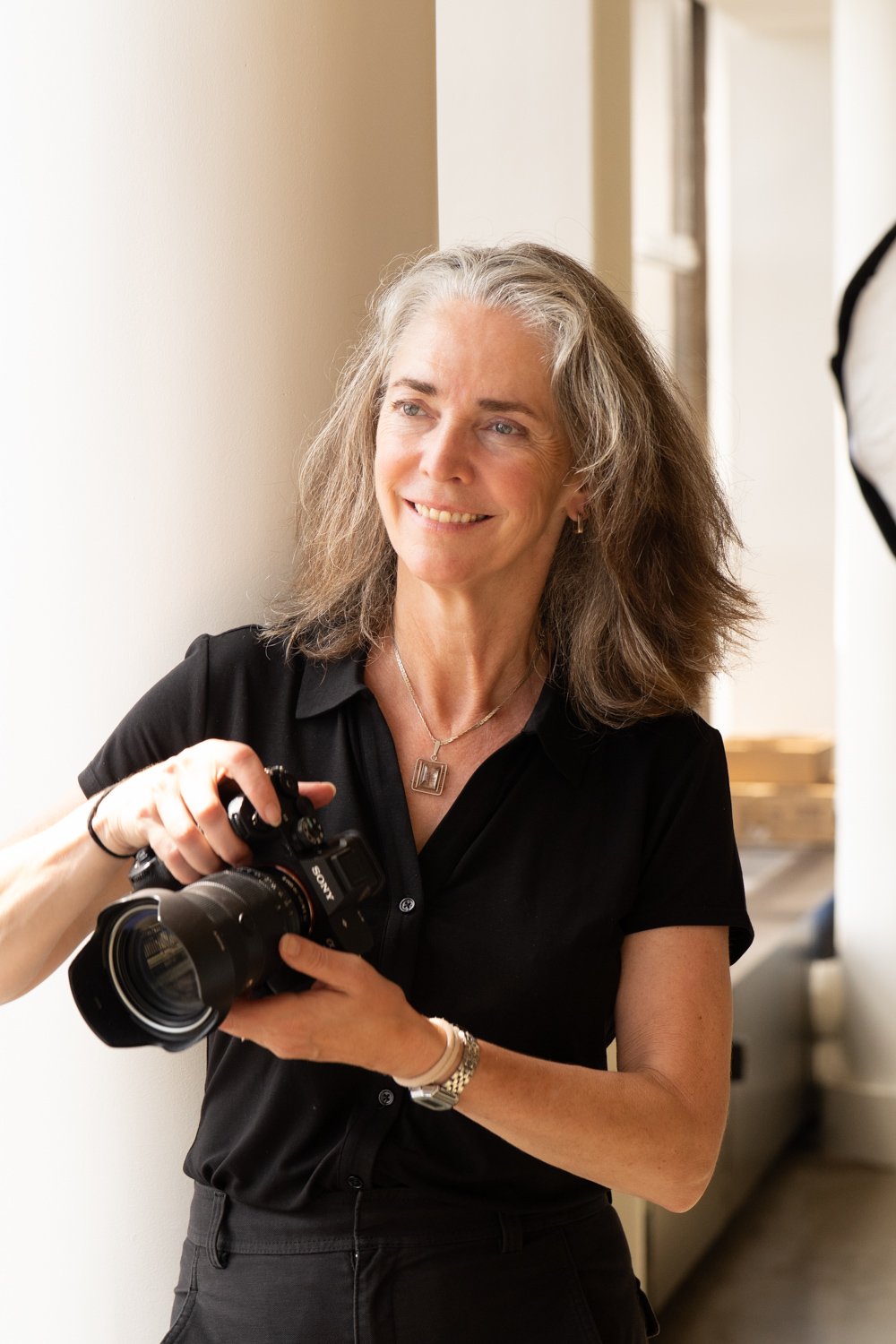 Photographer, Justine Cooper, holding a professional camera and warmly smiling, standing indoors at corporate offices near a window.