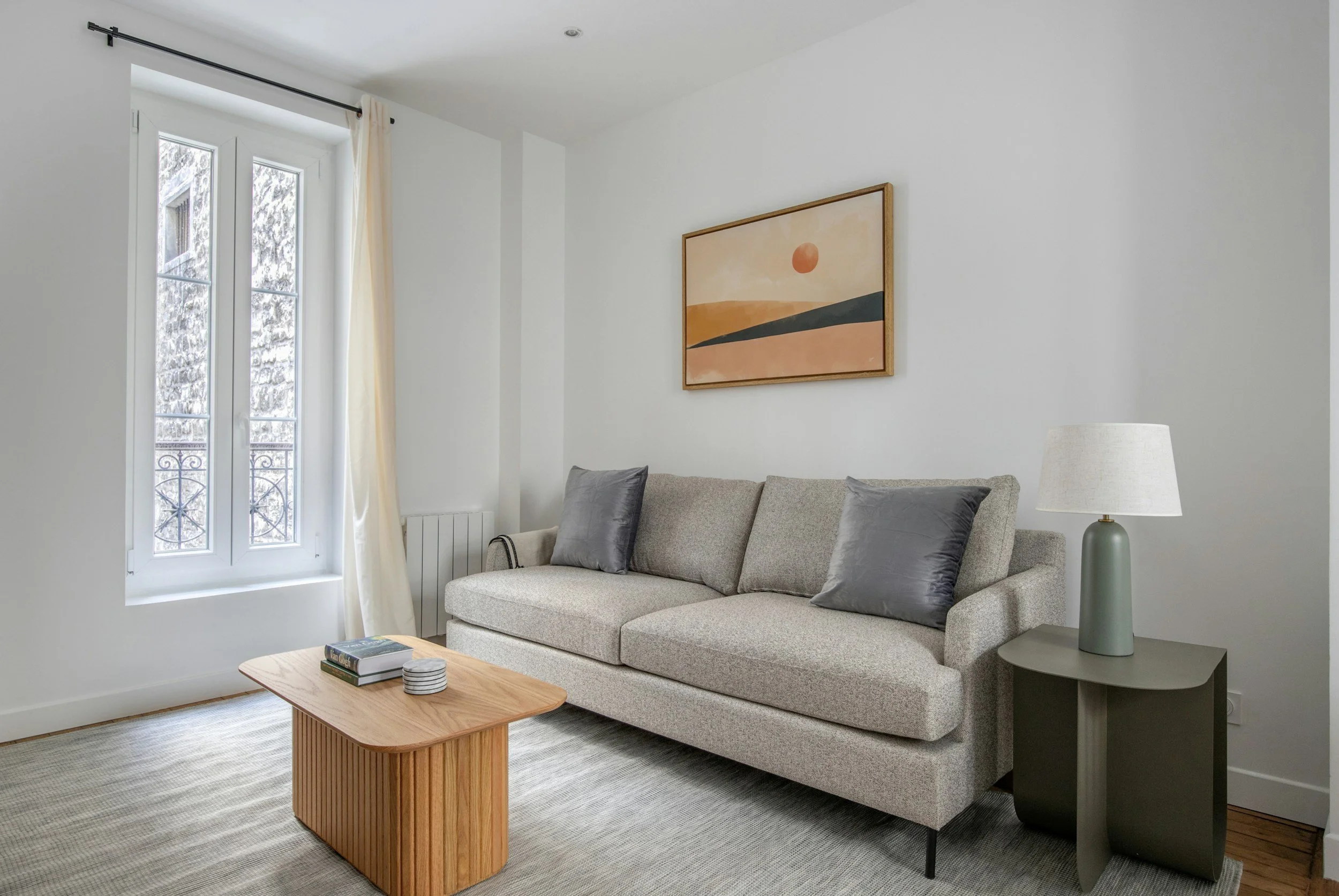 Living room with a large window, gray sofa with pillows, framed landscape art, side table with a lamp, and a coffee table with books and plates.