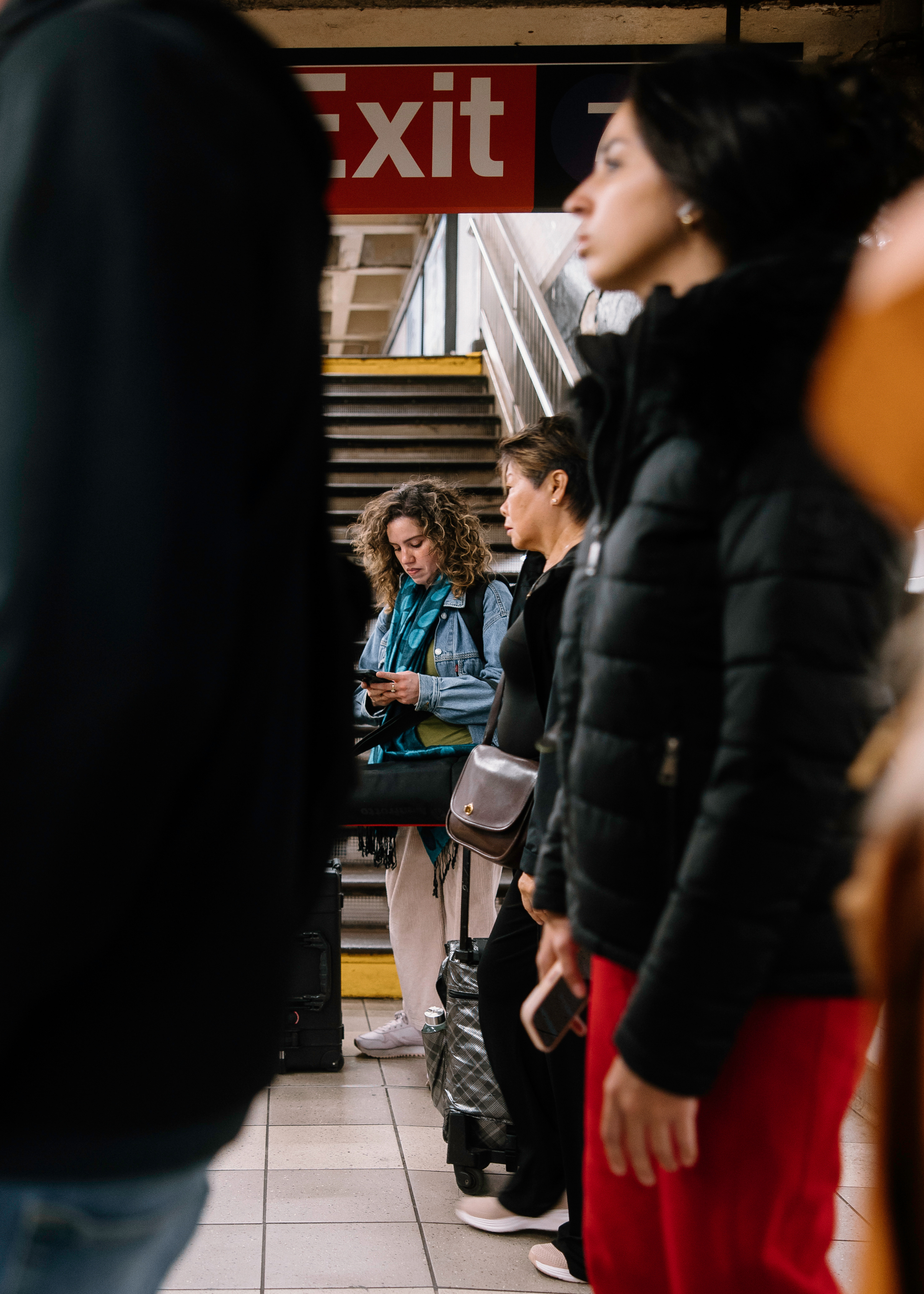 Street photography of people waiting in line at a New York City subway station with stairs and an exit sign in the background.