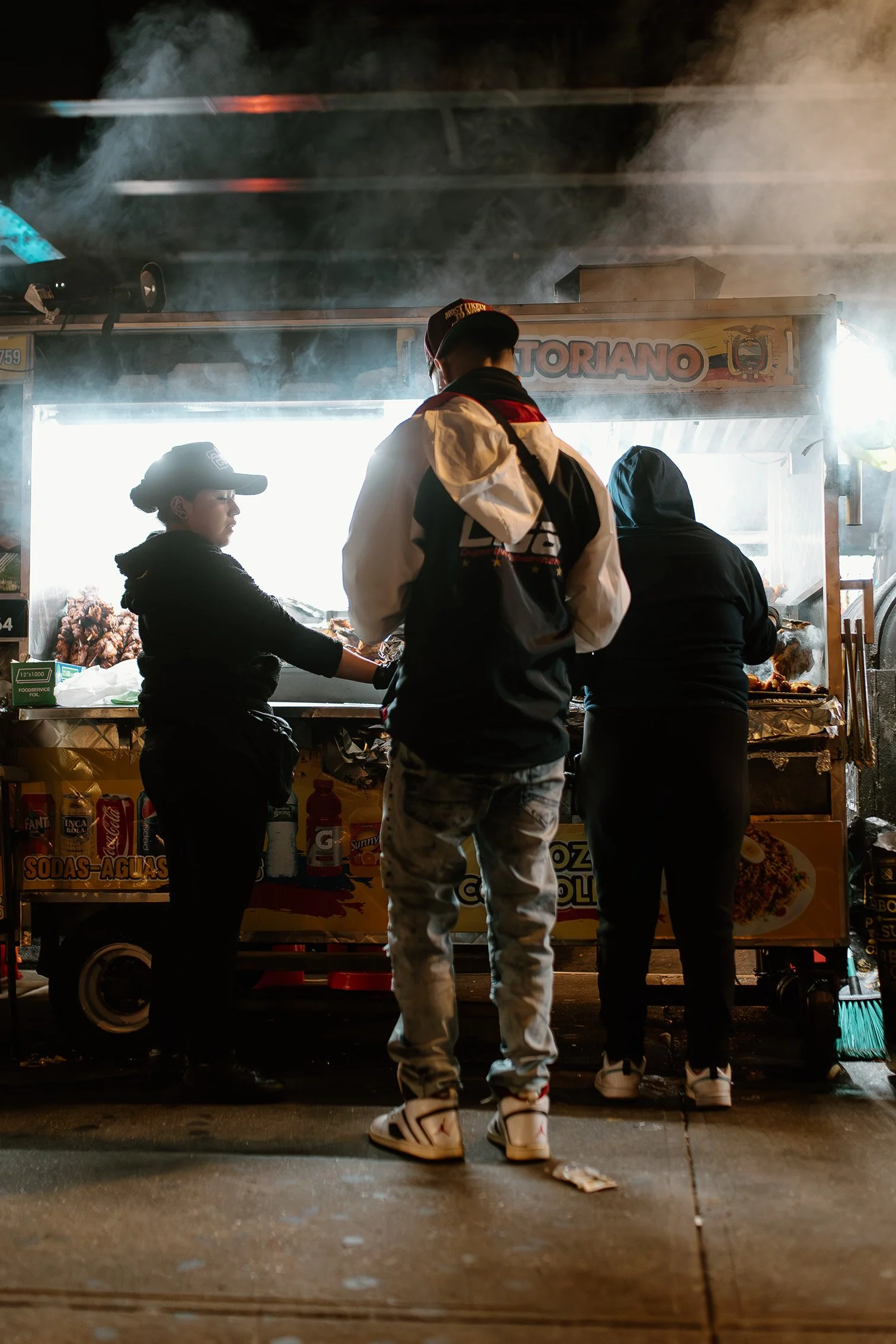 Night street photography of people gathered at an illuminated food stand with smoke rising into the city streets.