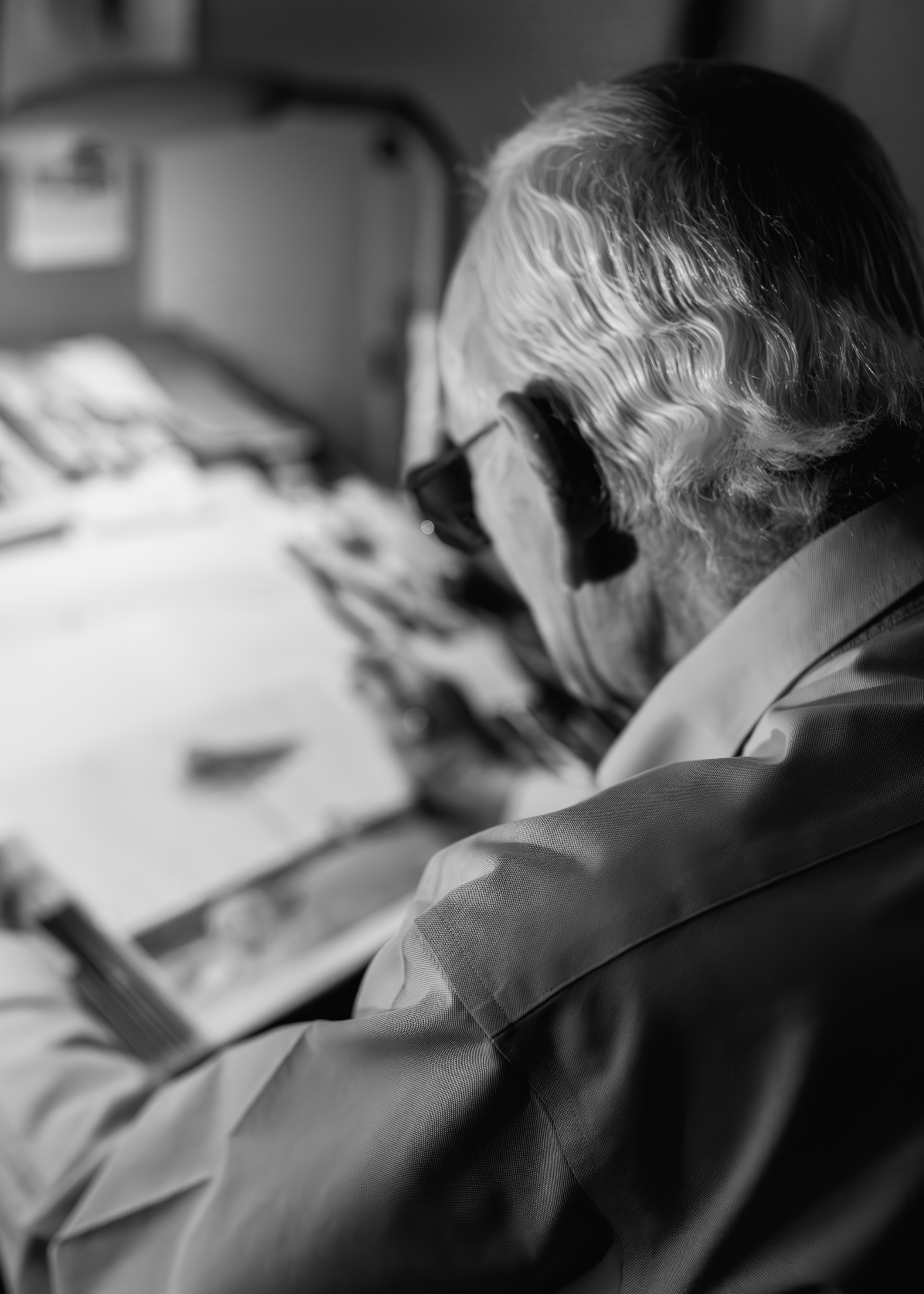 Close-up portrait of a person with wavy hair and glasses working at a desk with a computer in the background.