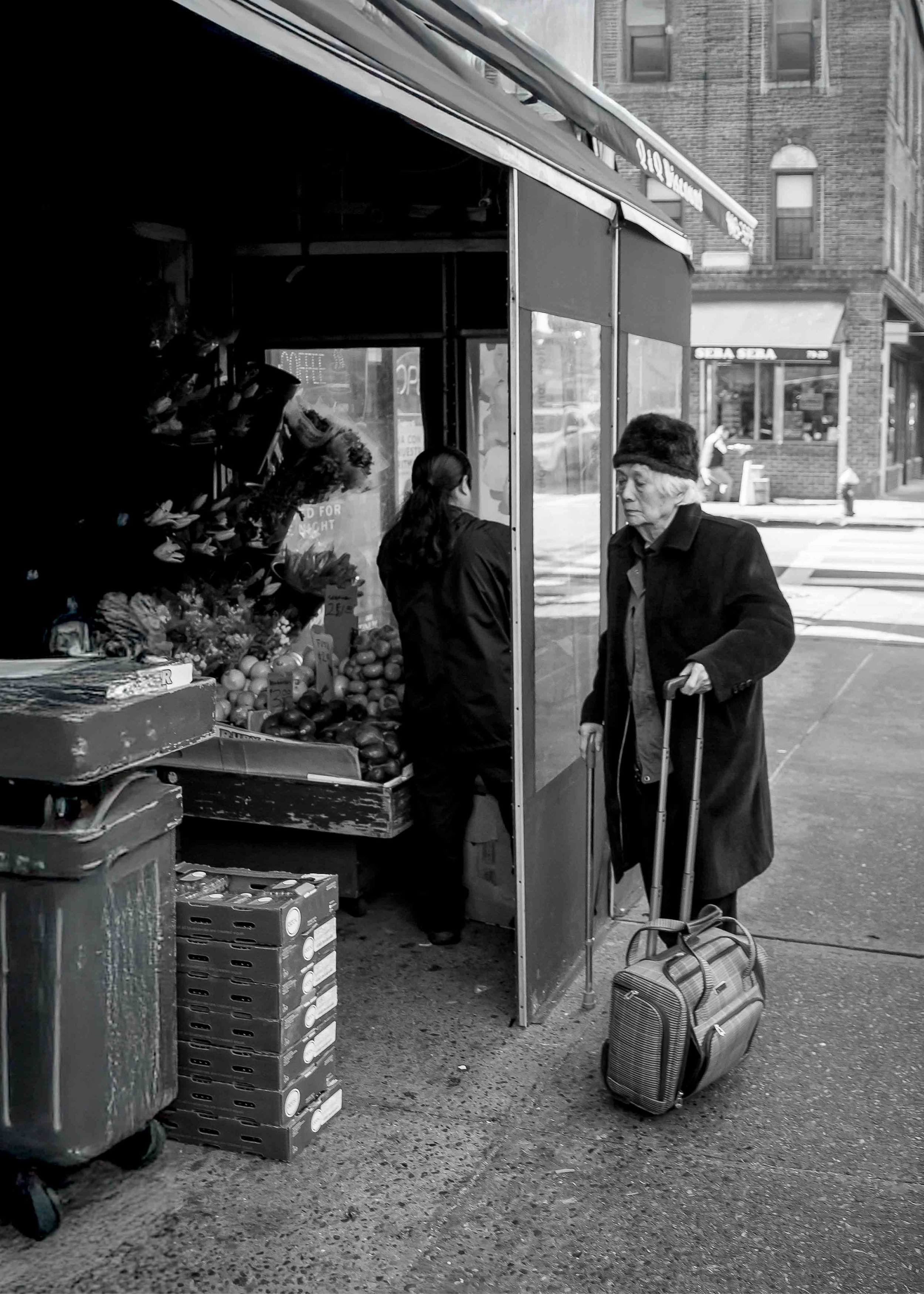 Documentary photograph of an elderly woman with luggage speaking to a vendor at a bustling city fruit and vegetable market.