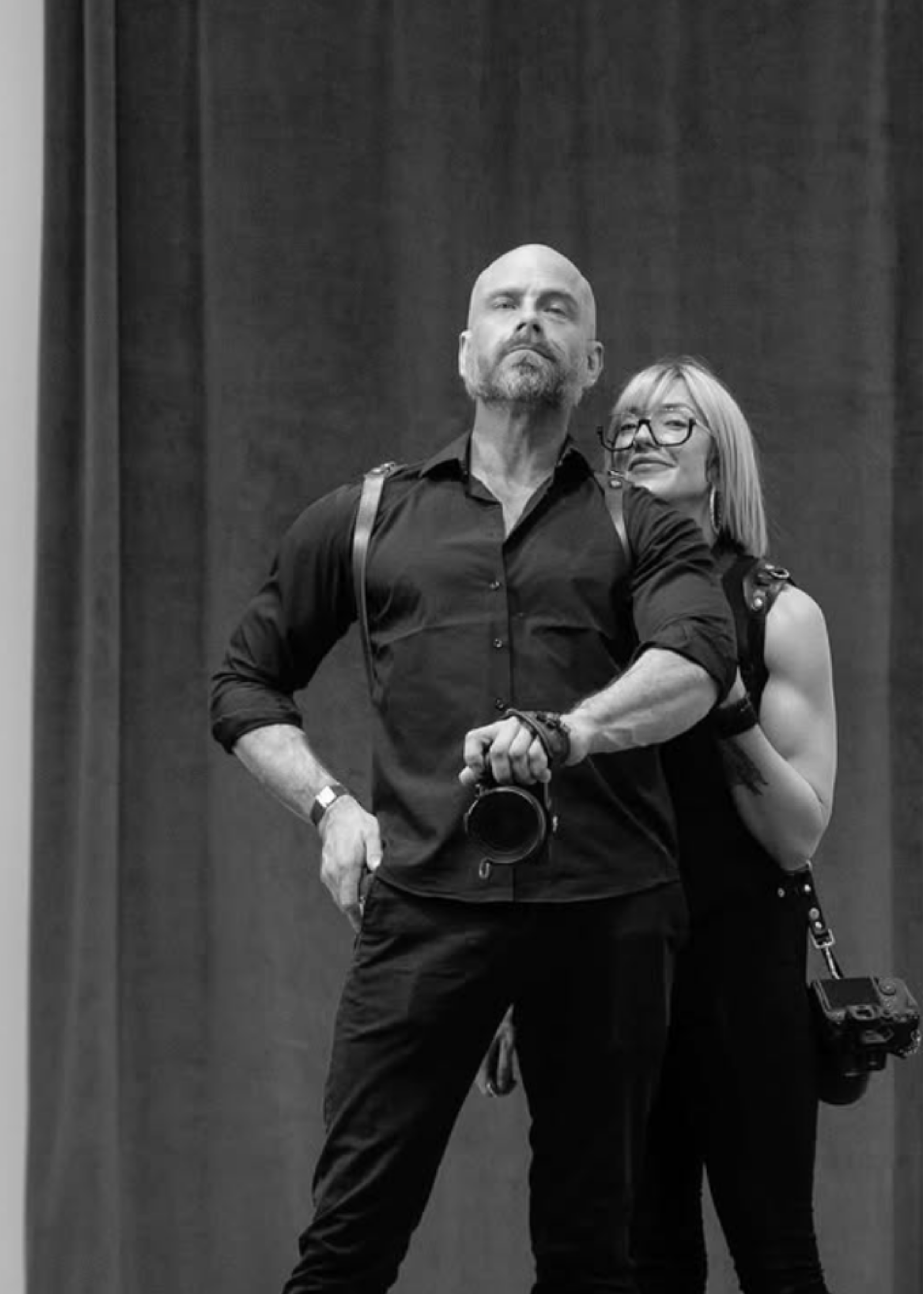 A black and white photograph of a man with a camera around his neck and a woman with glasses and light hair, both posing in front of a dark curtain.