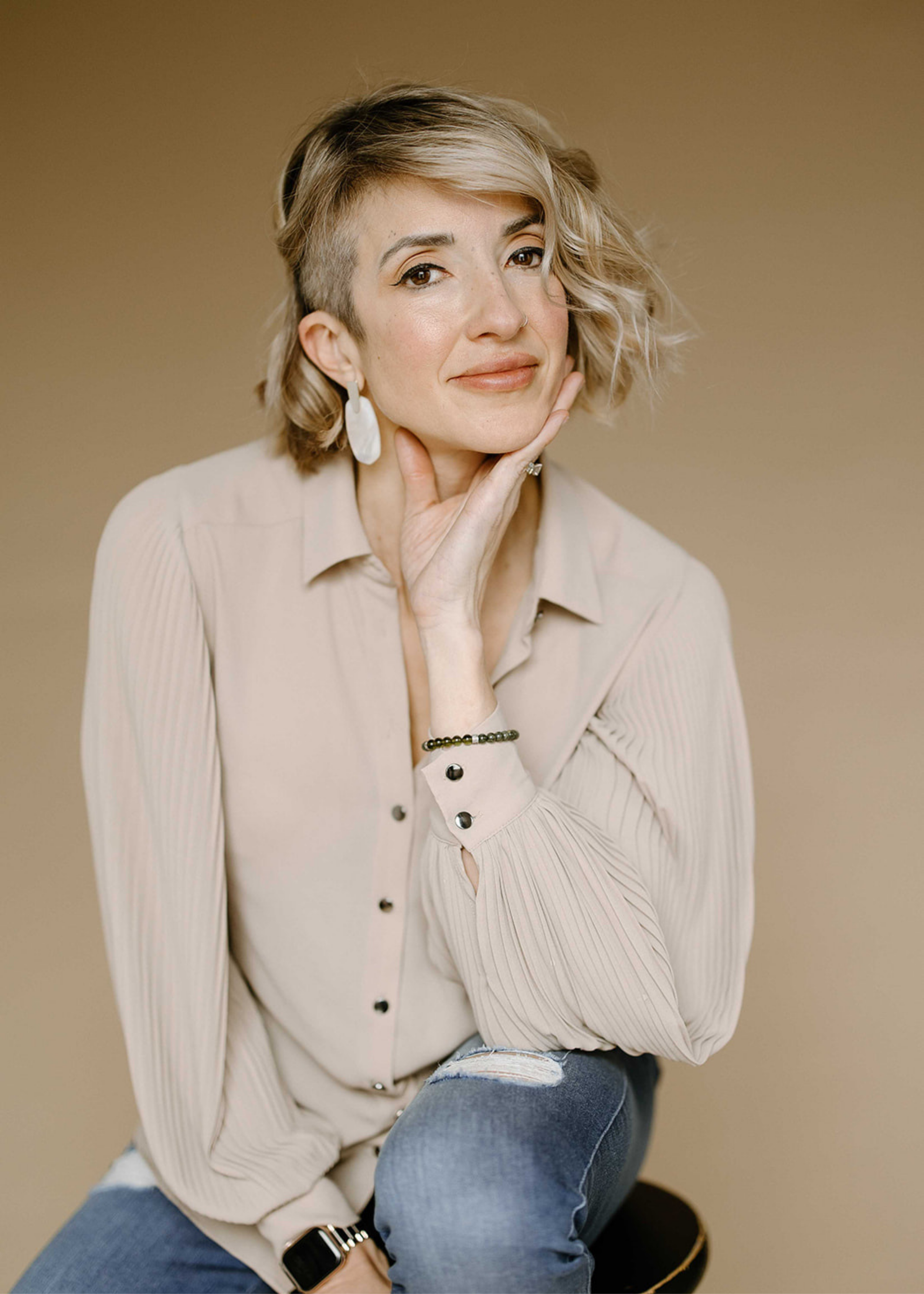 Portrait of Aperture & Atlas photographer Autumn with short wavy blonde hair, wearing a beige button-up shirt and ripped jeans against a neutral beige background.