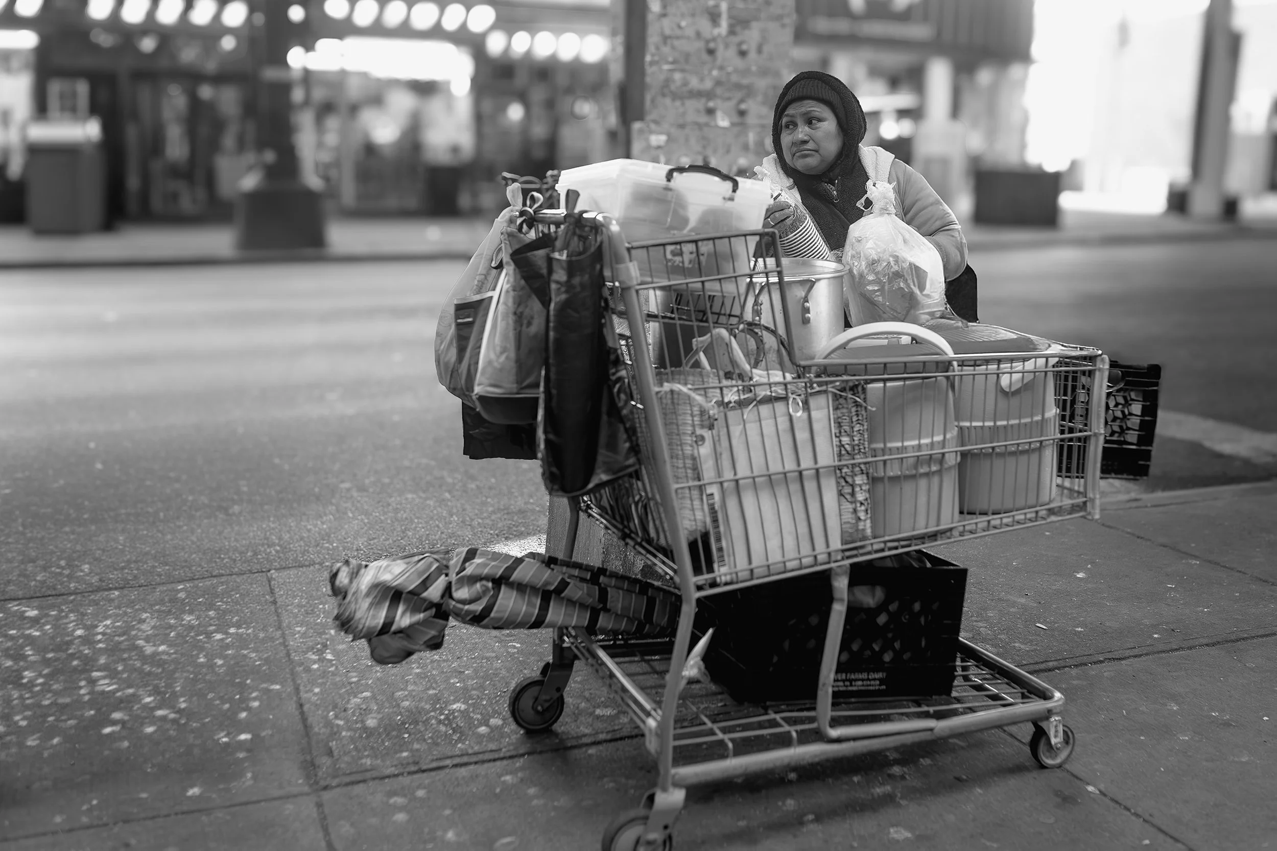 Documentary-style photograph of a woman standing behind a shopping cart filled with belongings on a city sidewalk at night.