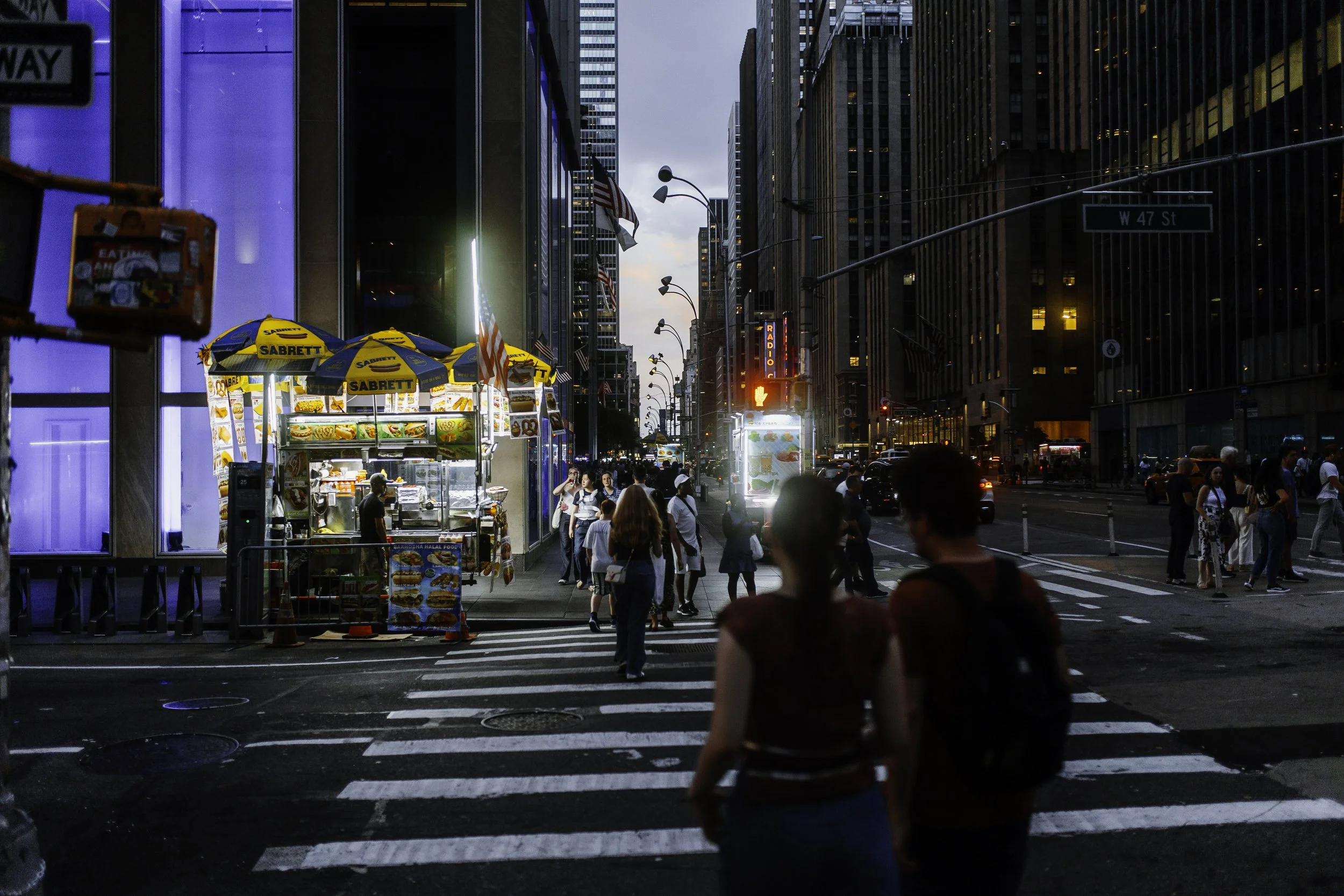 Street photography of pedestrians crossing a city intersection at dusk with a food cart, American flags, and surrounding skyscrapers.