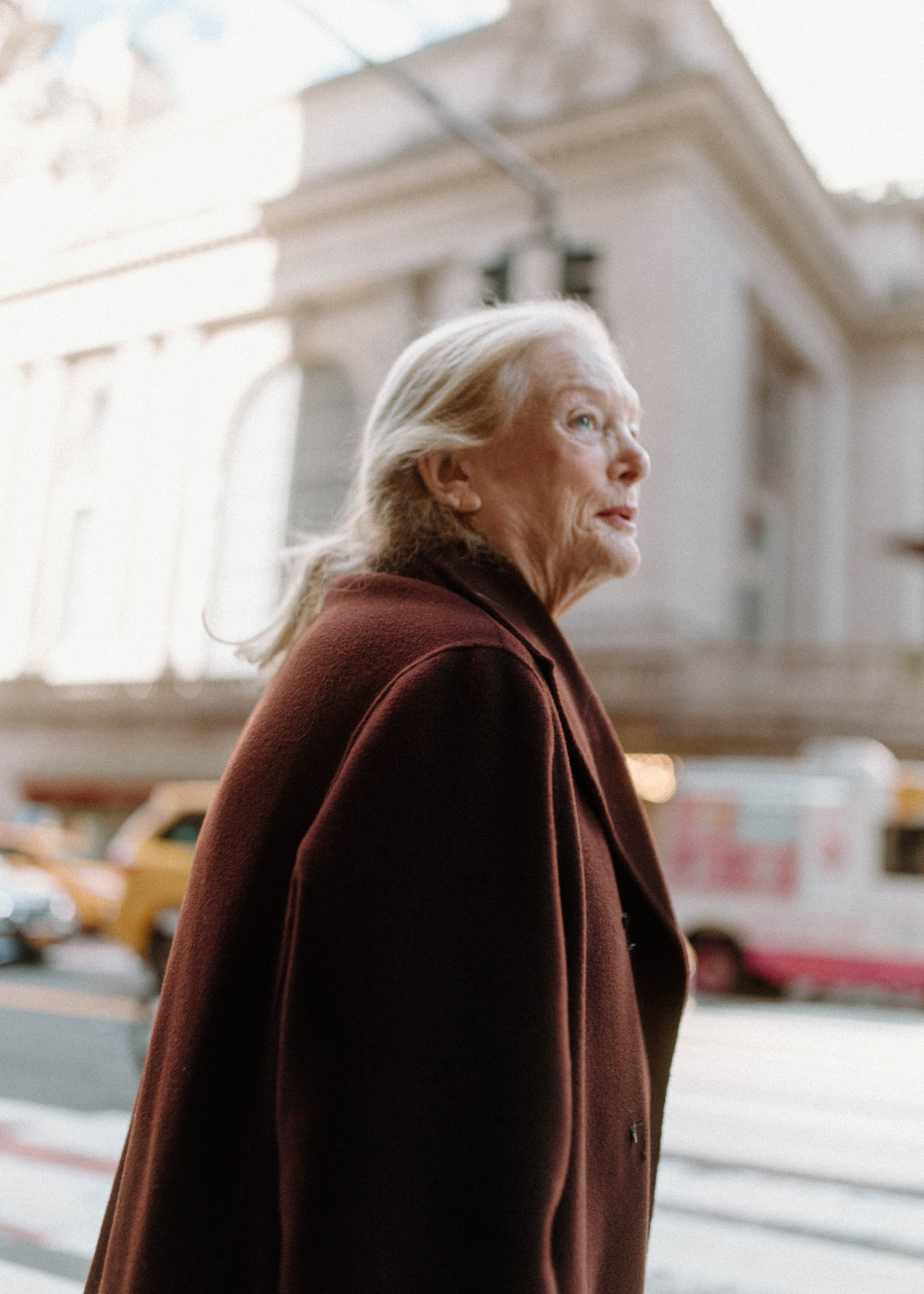 Documentary-style photograph of an elderly woman in a dark red coat walking along a city street with a blurred urban background.