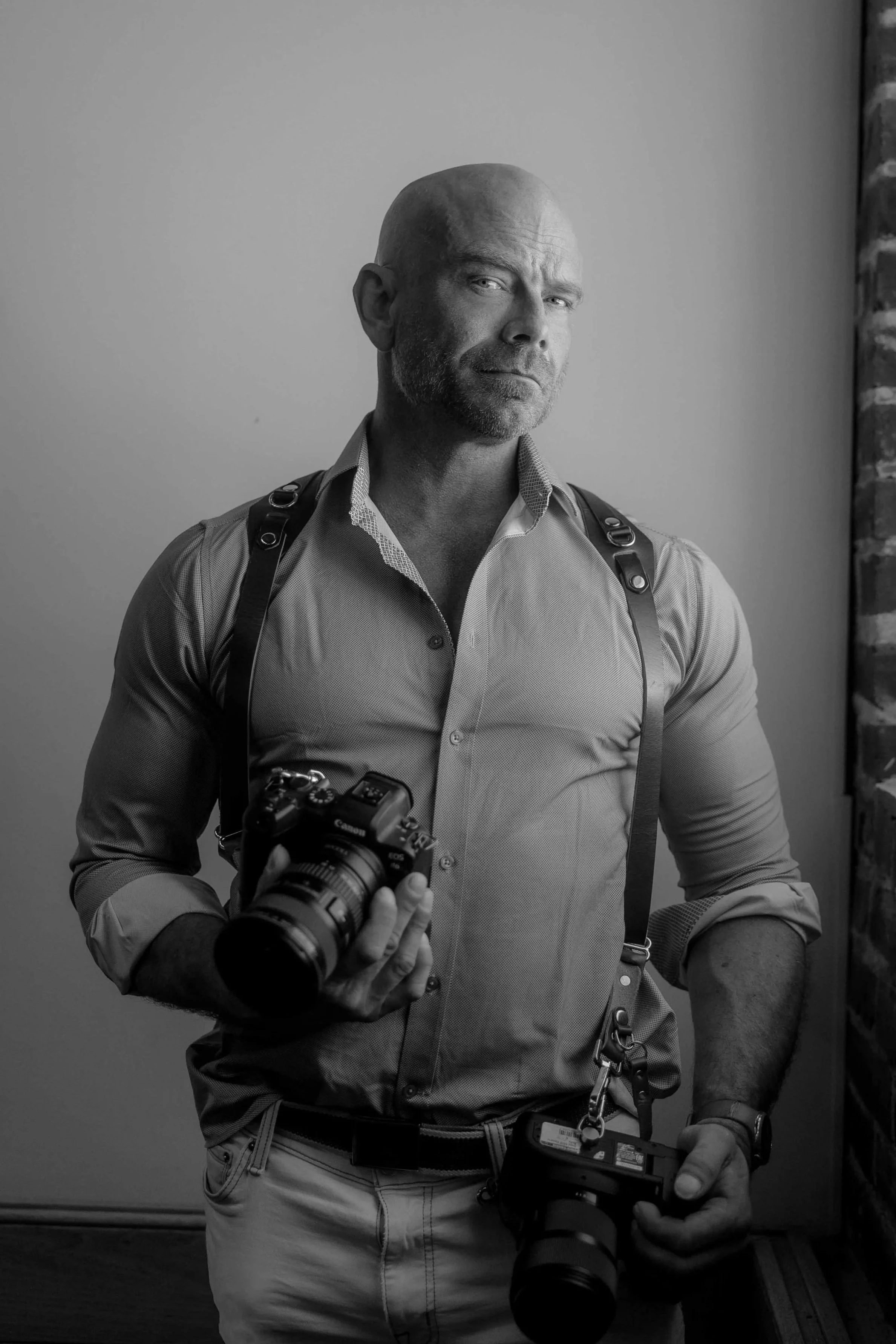 Black and white portrait of Aperture & Atlas photographer Stuart holding a professional camera and remote shutter release against a textured studio wall.