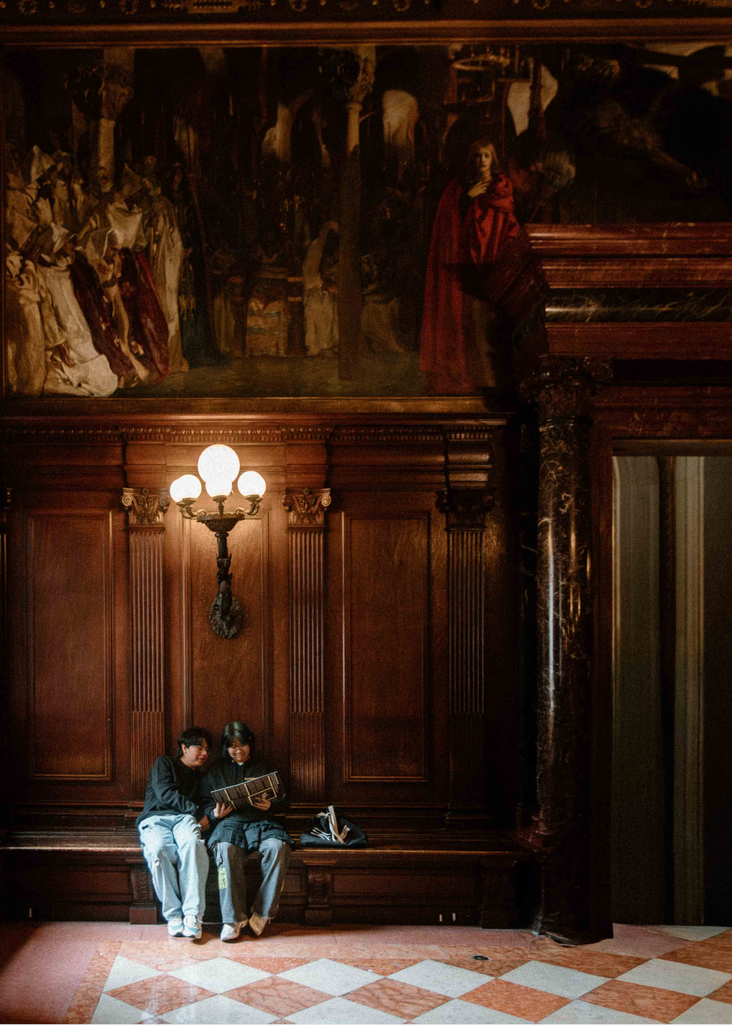 Lifestyle photograph of two women sitting on a wooden bench reading a book together in an elegant hallway with marble floors and wood-paneled walls.