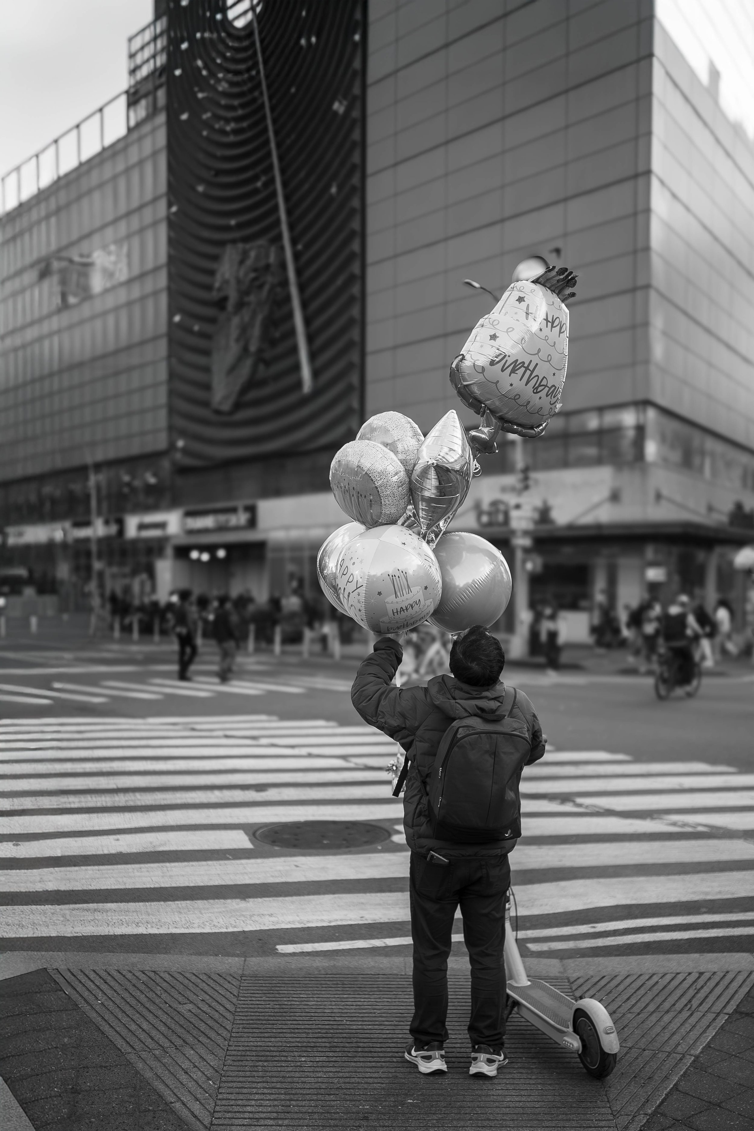 Street photograph of a person with a backpack carrying balloons while crossing a city street in front of a modern building.