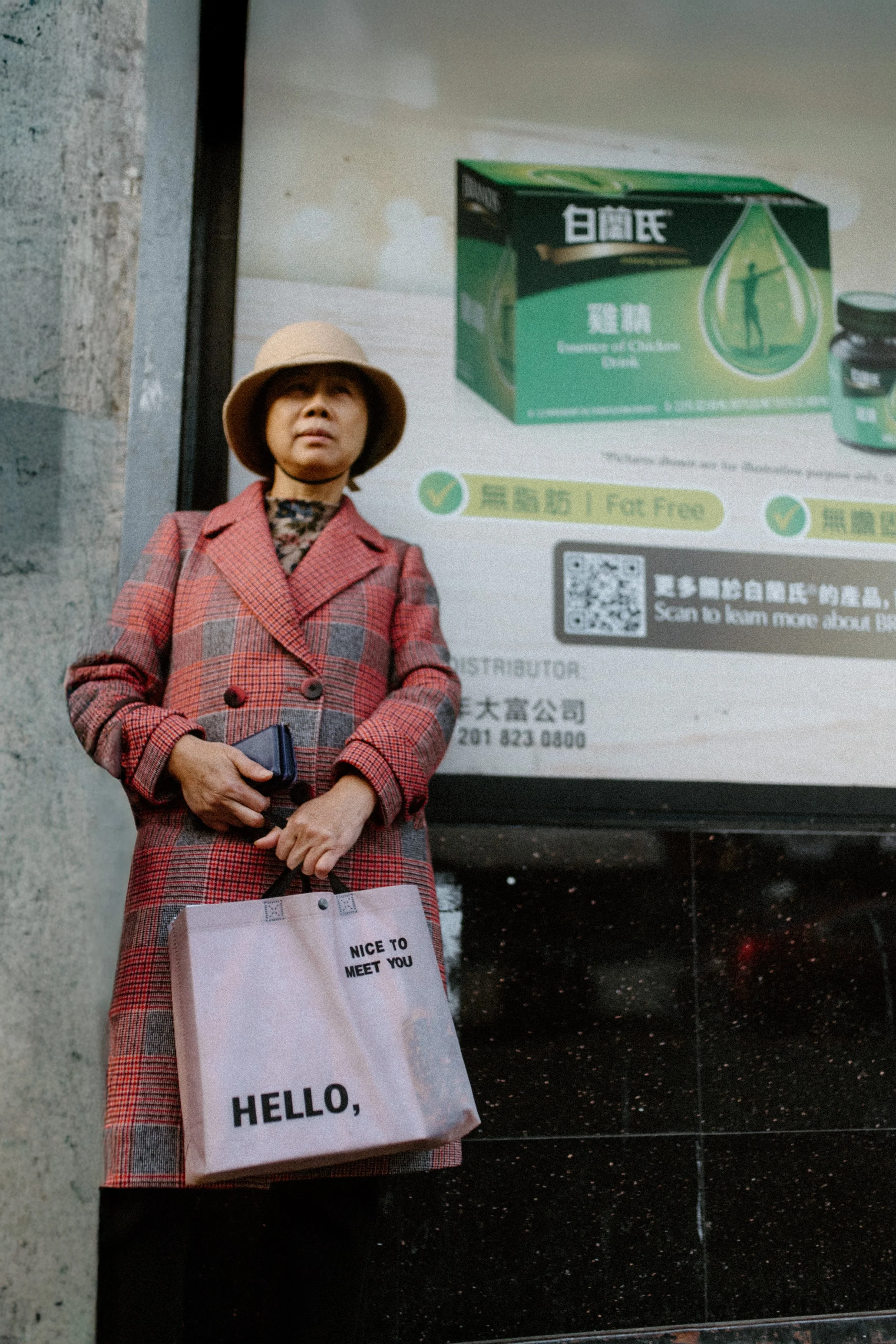 Street photograph of a woman in a red plaid coat standing in front of a colorful city advertisement with English and Chinese text.
