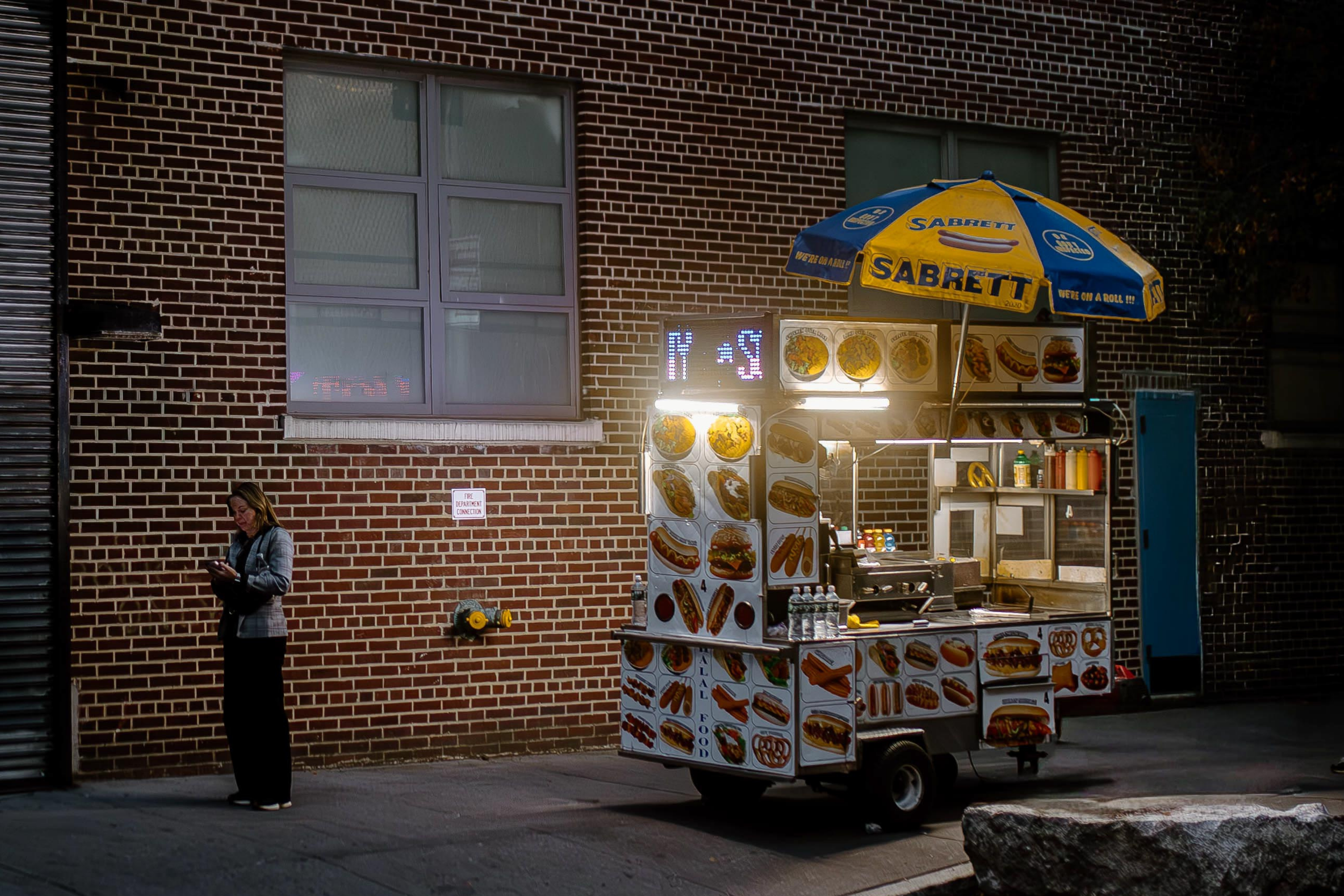 Street photograph of a colorful hot dog stand outside a brick building with a woman checking her phone on the sidewalk.