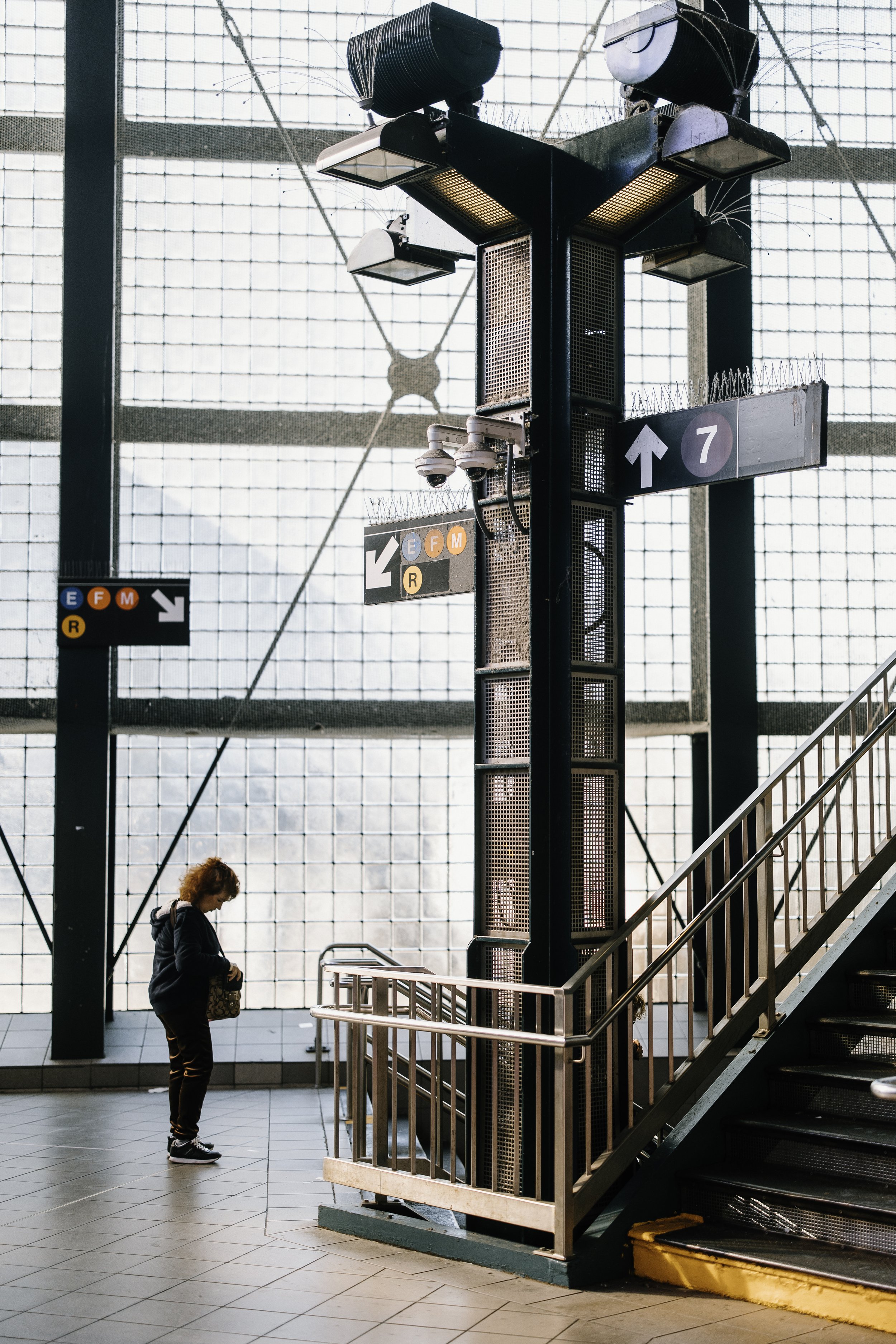 Street photograph of a woman waiting on a subway platform while looking at her phone beneath industrial lighting.