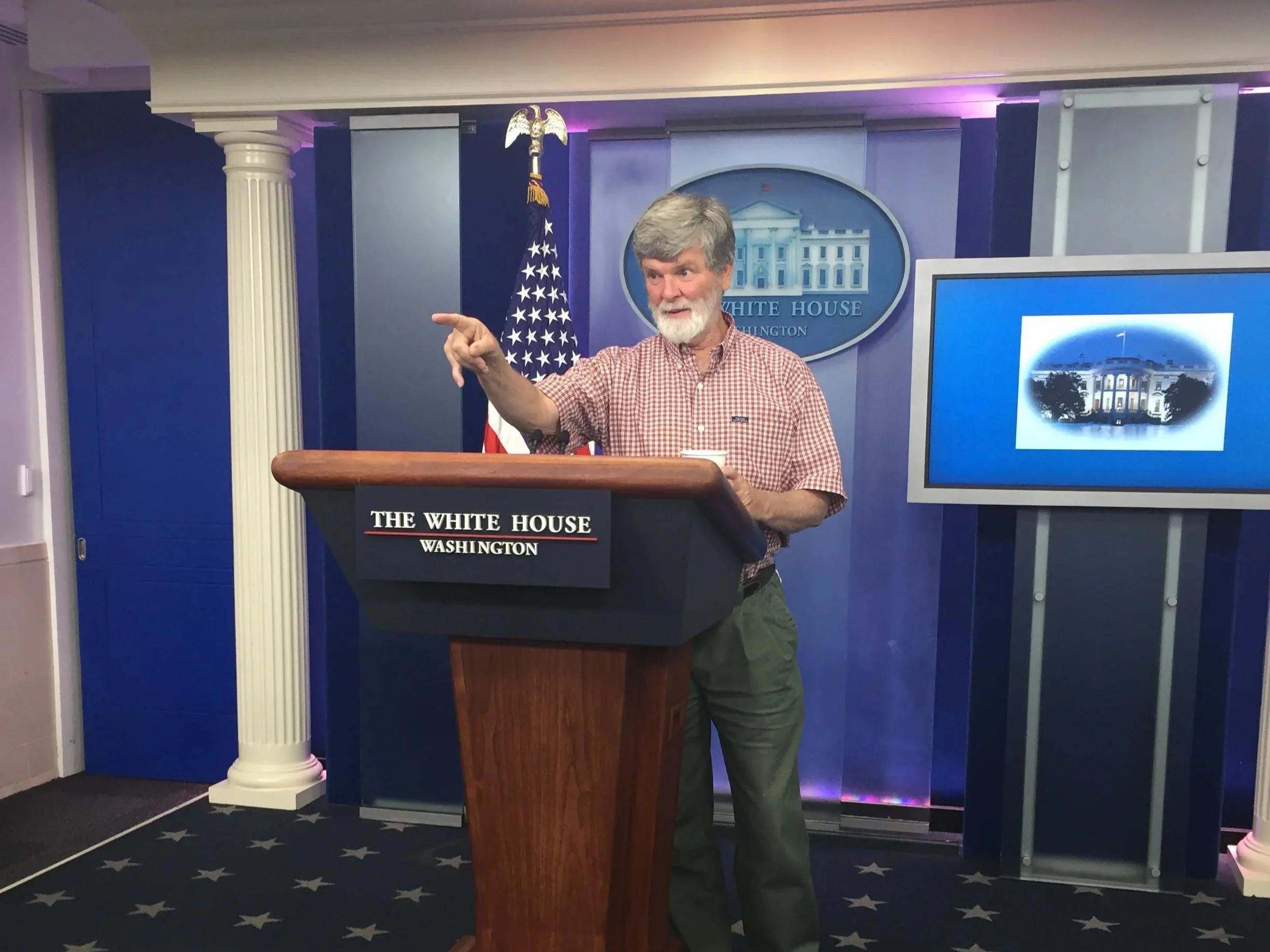 A man with gray hair and a beard wearing a red checkered shirt standing at a podium in the White House press briefing room, pointing to the side.