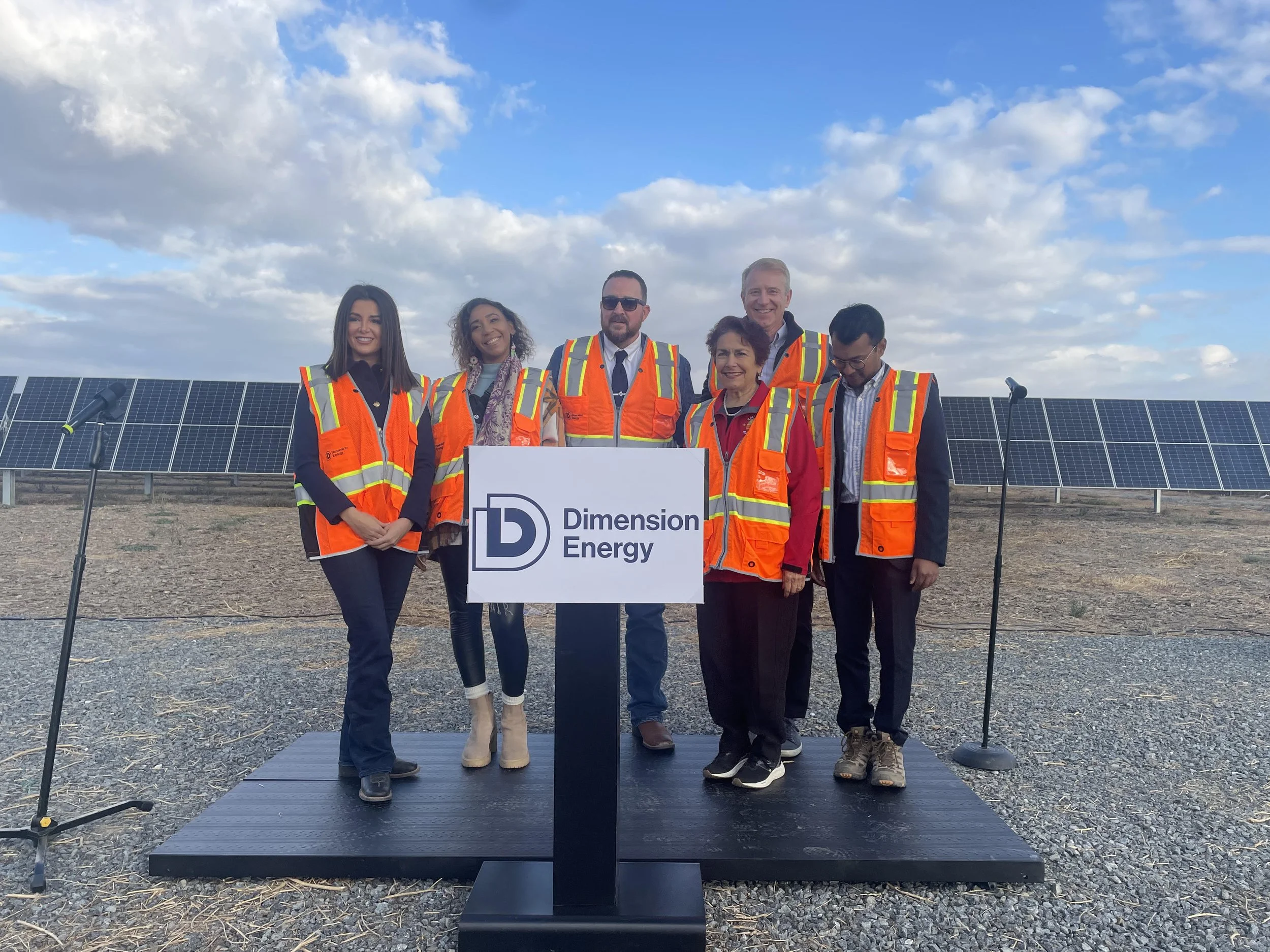 Group of six people standing outdoors in front of solar panels, wearing safety vests, at a presentation or event for Dimension Energy.