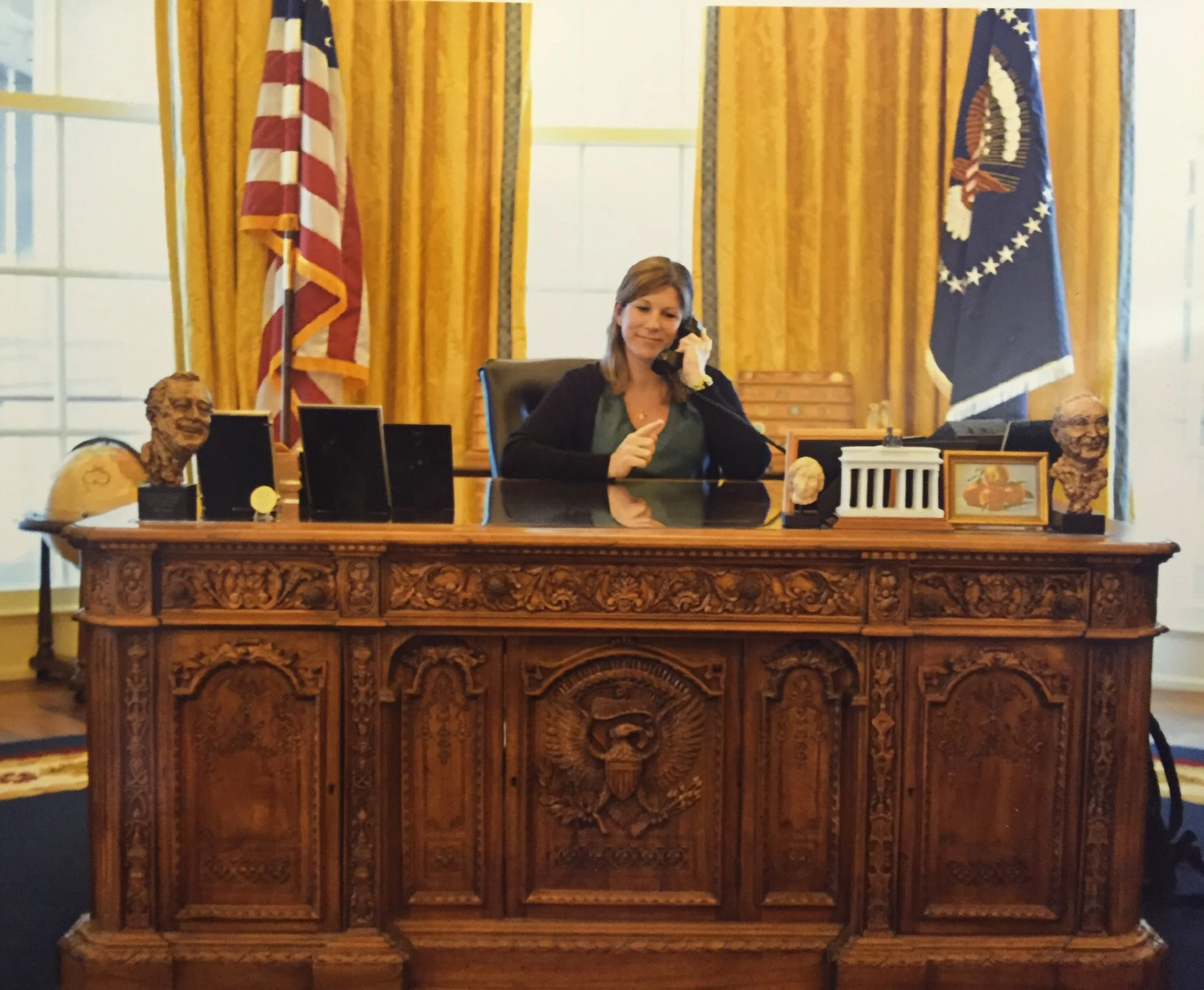 A woman sitting at a large ornate wooden desk in a room with yellow curtains and flags of the United States and the President of the United States, holding a phone receiver to her ear, surrounded by framed photographs and busts.