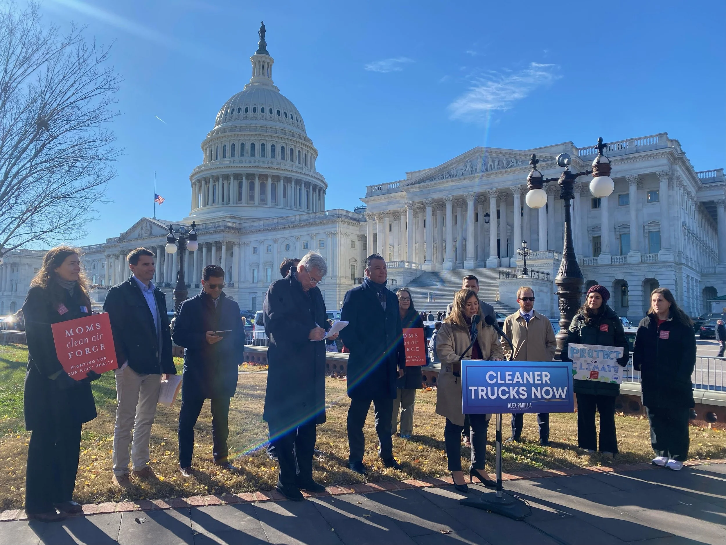 A group of people protesting in front of the United States Capitol building, holding signs related to climate change and cleaner trucks, on a sunny day with clear blue skies.