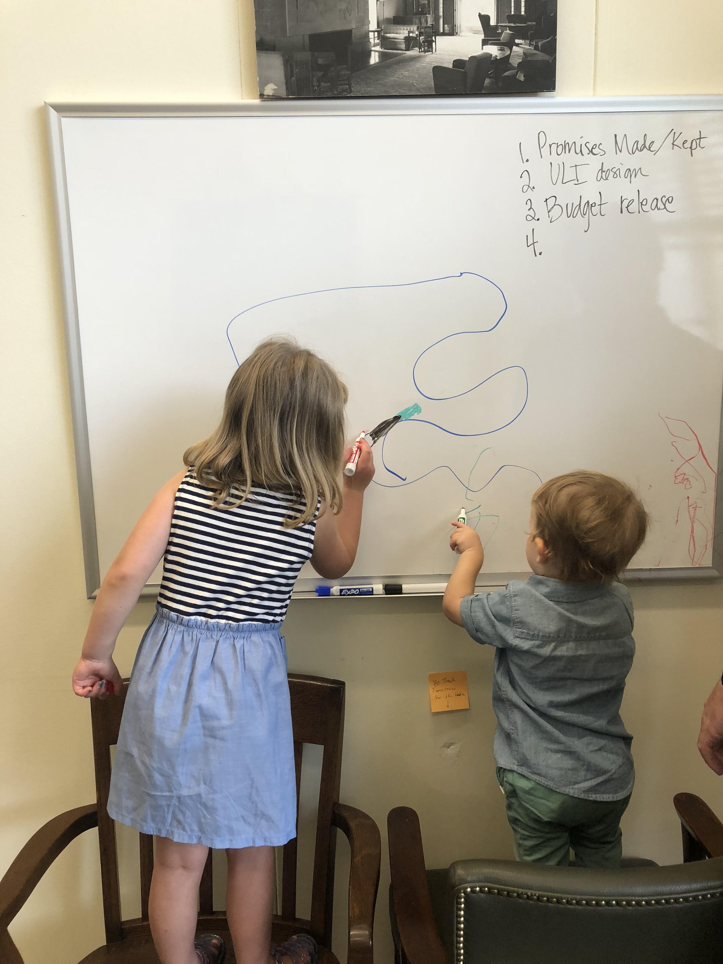 Two children drawing on a whiteboard, with chairs surrounding them, in a room that appears to be a classroom or library, with a picture of a living room hanging on the wall.