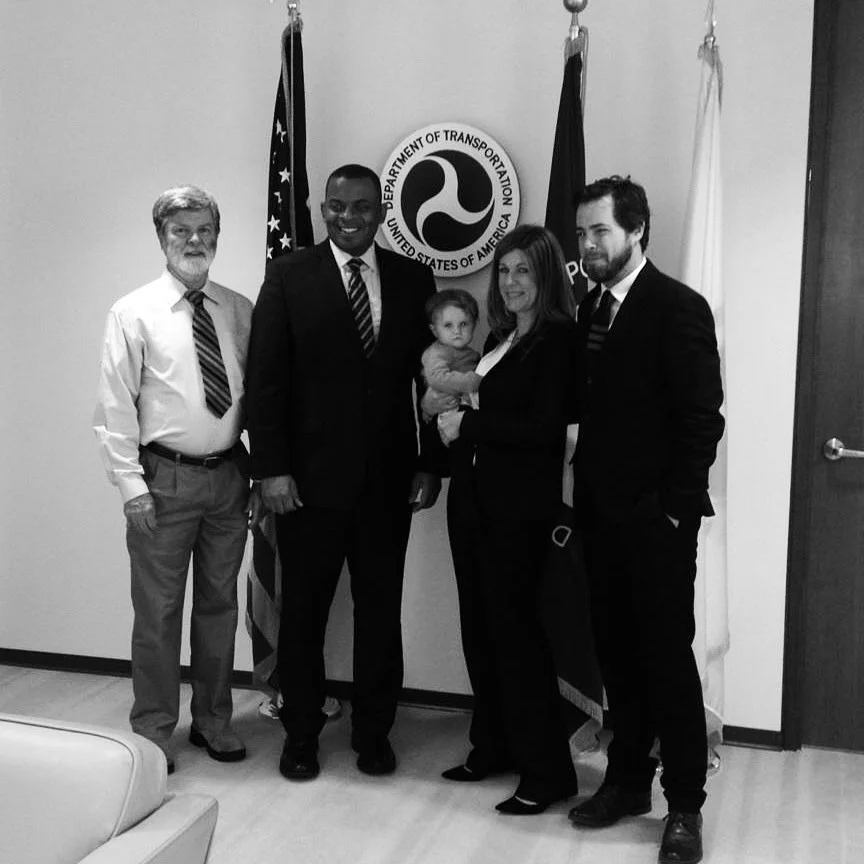 Group of five adults and one child standing in front of a sign for the U.S. Department of Transportation, with American and departmental flags, in an office setting.