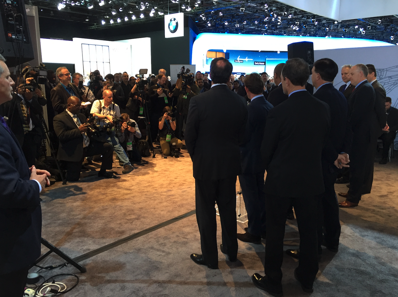 Business executives speaking at a press conference or event, with numerous photographers and journalists capturing the moment, surrounded by a display at an auto or technology trade show.