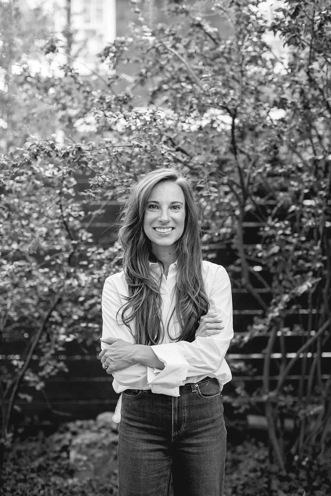 A woman with long wavy hair, smiling, wearing a white shirt and dark jeans, standing outdoors in front of trees and bushes.