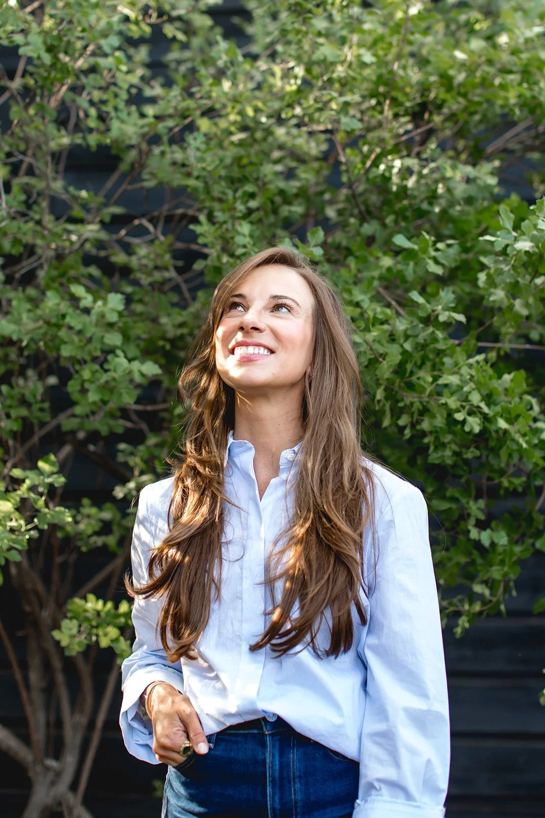A woman with long, wavy brown hair, wearing a white button-up shirt and blue jeans, standing outdoors in front of green foliage, smiling and looking upward.