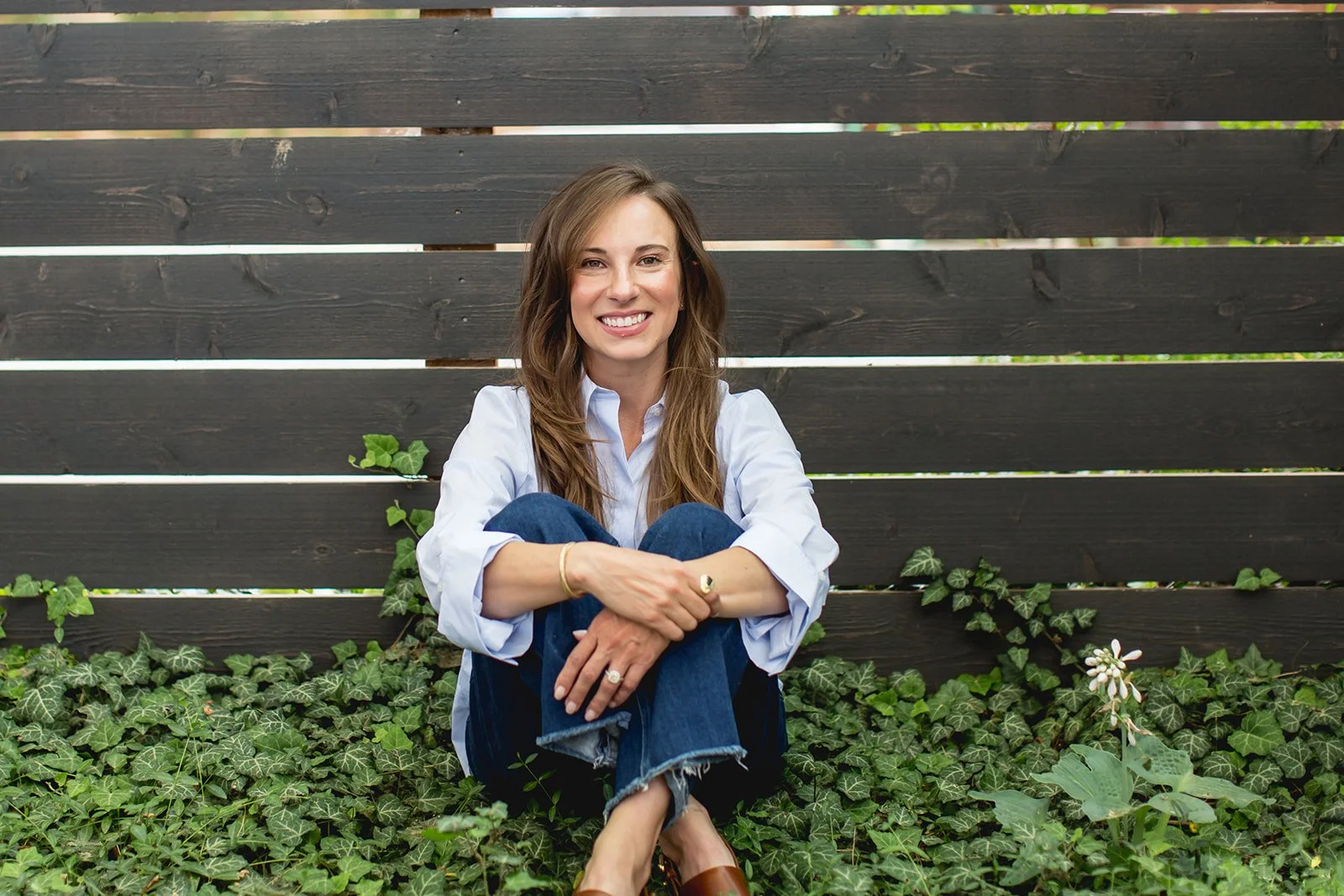 A woman sitting cross-legged on the ground with green ivy and plants, smiling, in front of a dark wooden fence with some plants growing around her.