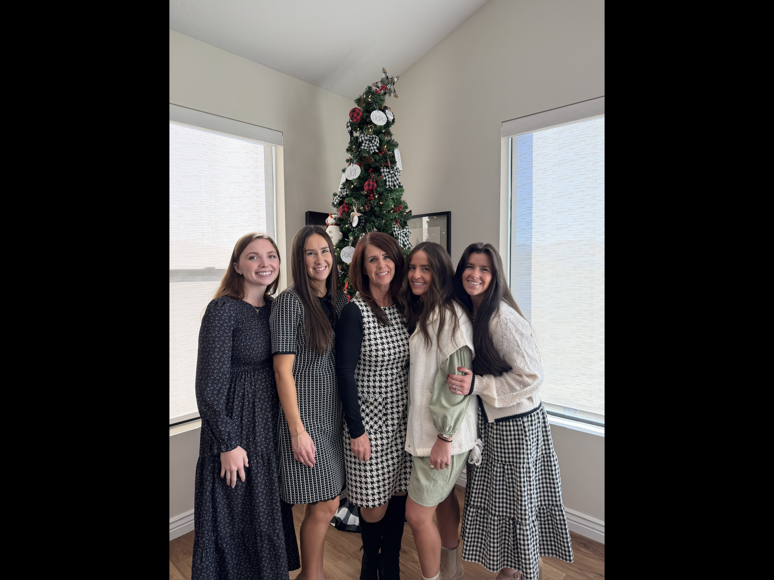 A group of five women smiling and standing in front of a decorated Christmas tree in a bright room with large windows.