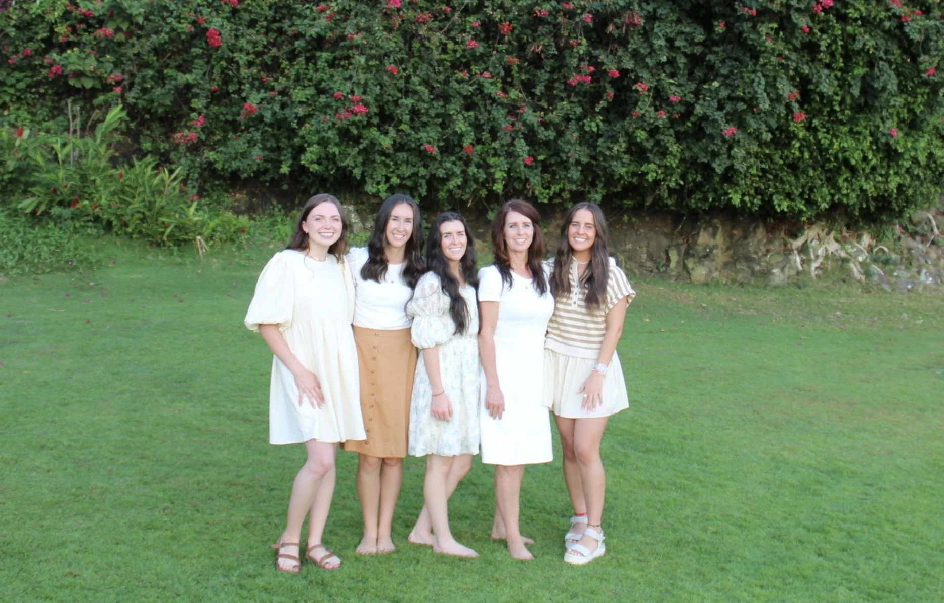 Five women standing on a grassy lawn in front of a bush with pink flowers, smiling at the camera.