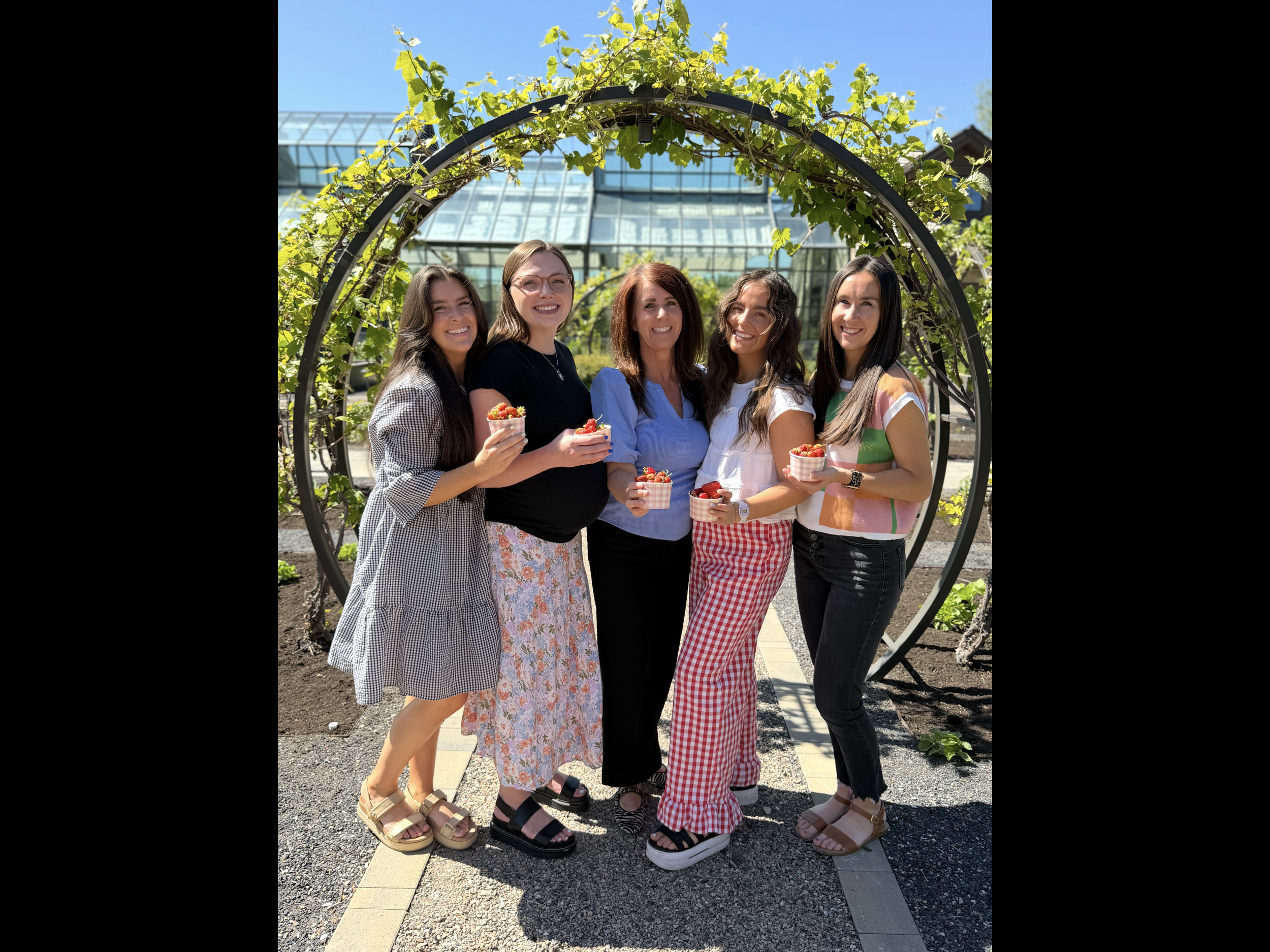 Five women standing together outdoors, smiling, holding small bowls of strawberries, with a greenhouse and blue sky in the background.