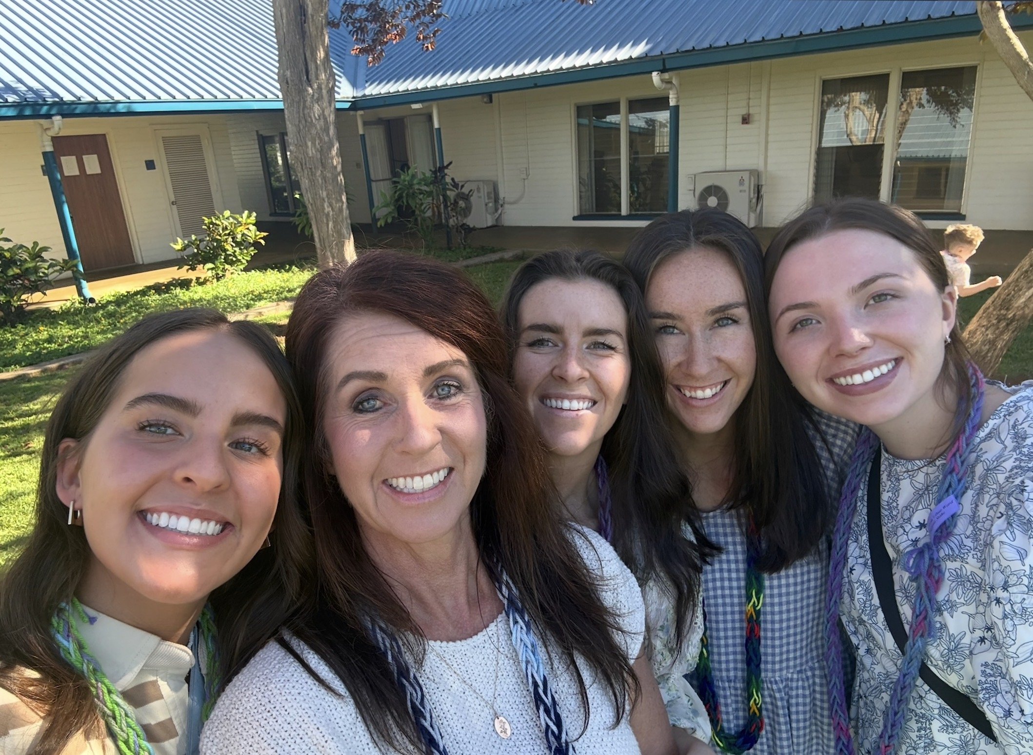 Group of five women smiling closely together outdoors in front of a building with trees and bushes.