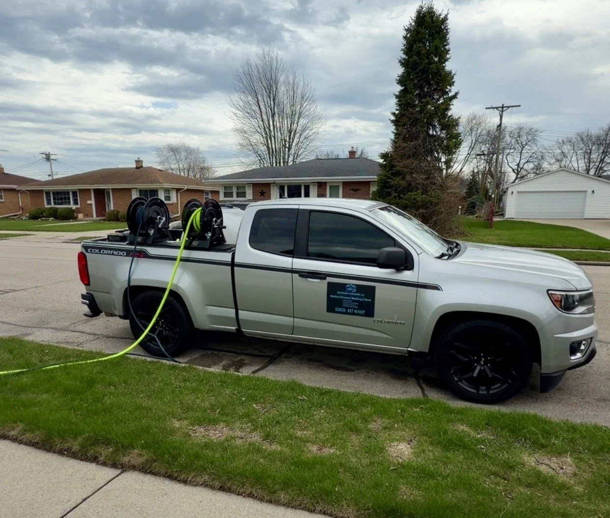A silver pickup truck parked on a residential street with a hose reel on the back, connected to a water source.