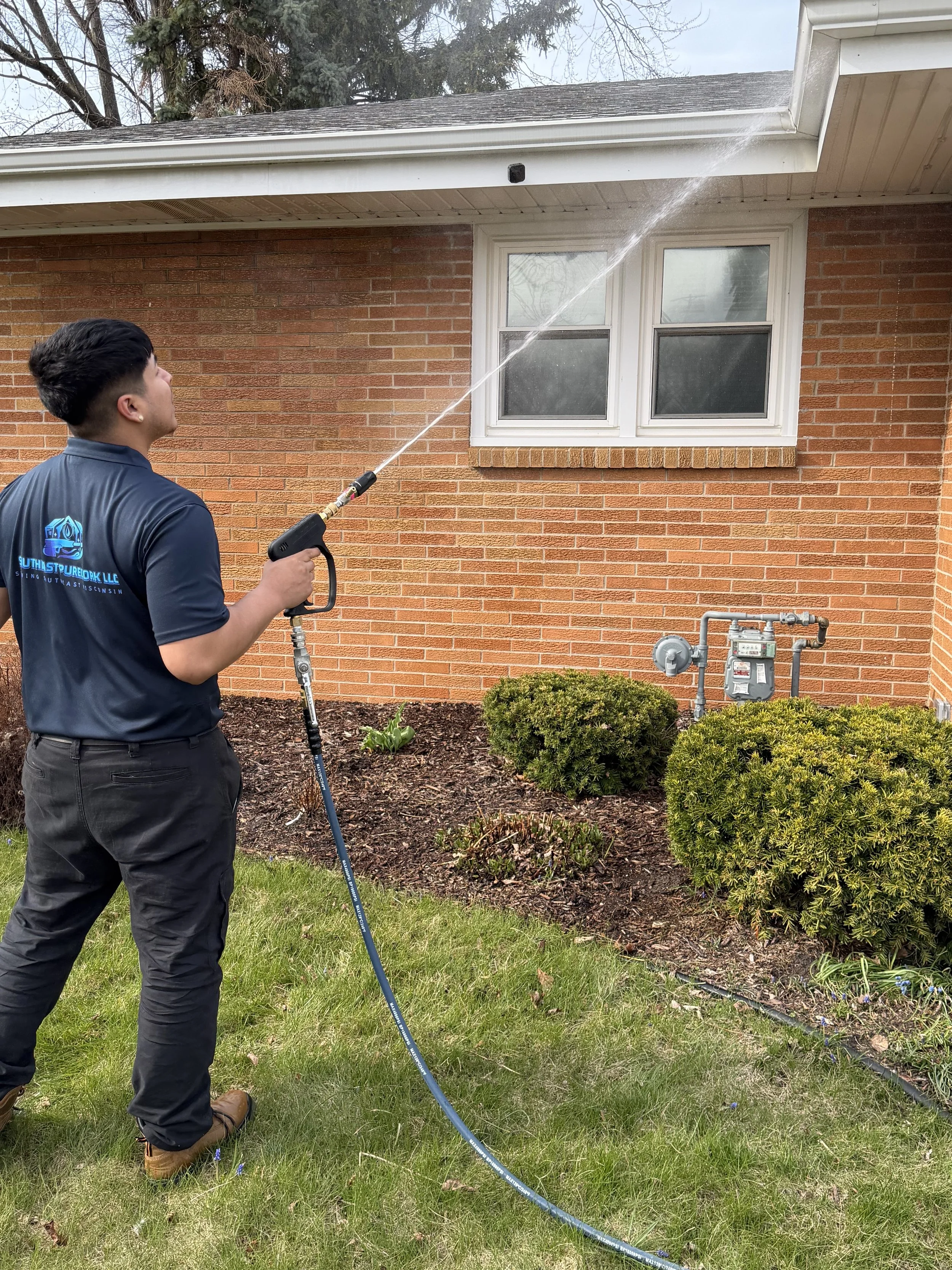 A man wearing a navy blue shirt and black pants is using a pressure washer to clean the exterior of a brick house. He is standing on grass, and there are bushes and a garden bed with plants nearby. The house has two windows and a roof with gray shingles.