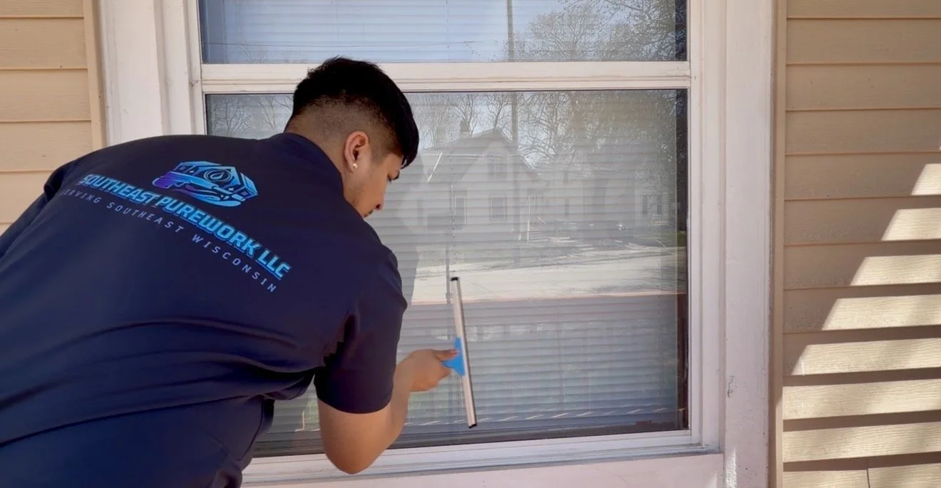 A person in a dark shirt with blue lettering is cleaning a window with a squeegee outside a house.