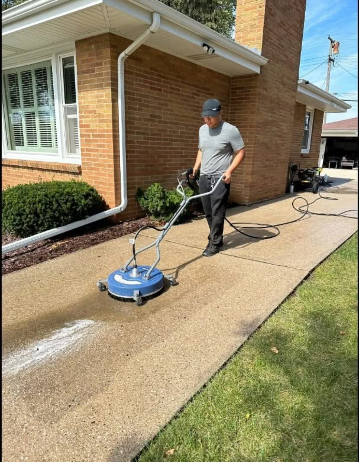 A person using a floor scrubber on a concrete sidewalk outside a brick house, with green grass and bushes nearby.
