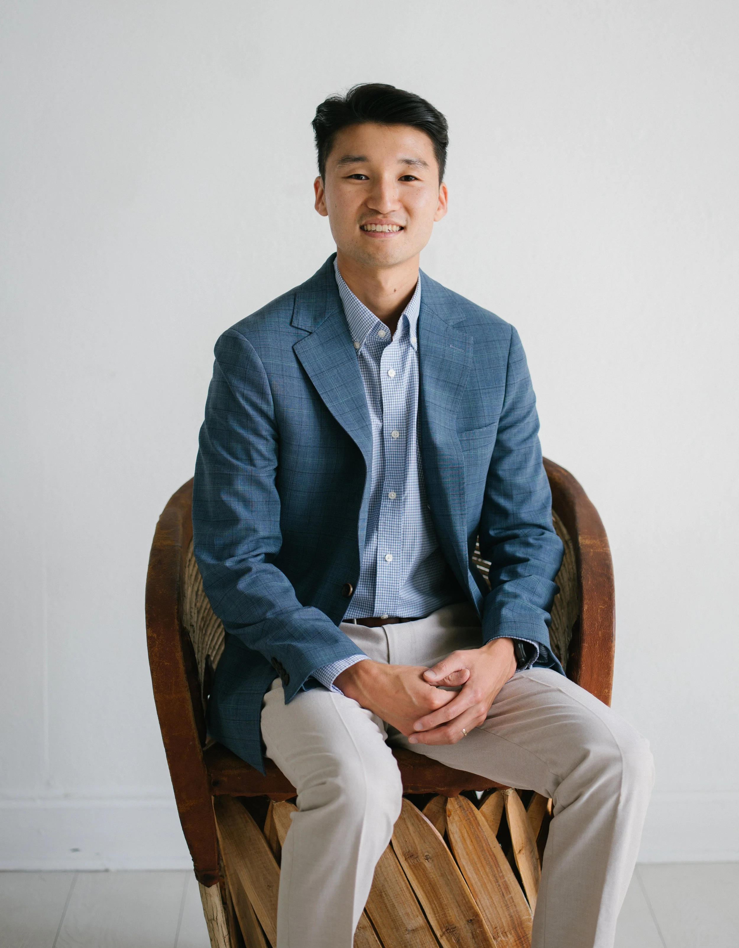A young Asian man with black hair sitting on a wooden chair against a plain white wall, wearing a blue checked blazer and beige pants, smiling at the camera.
