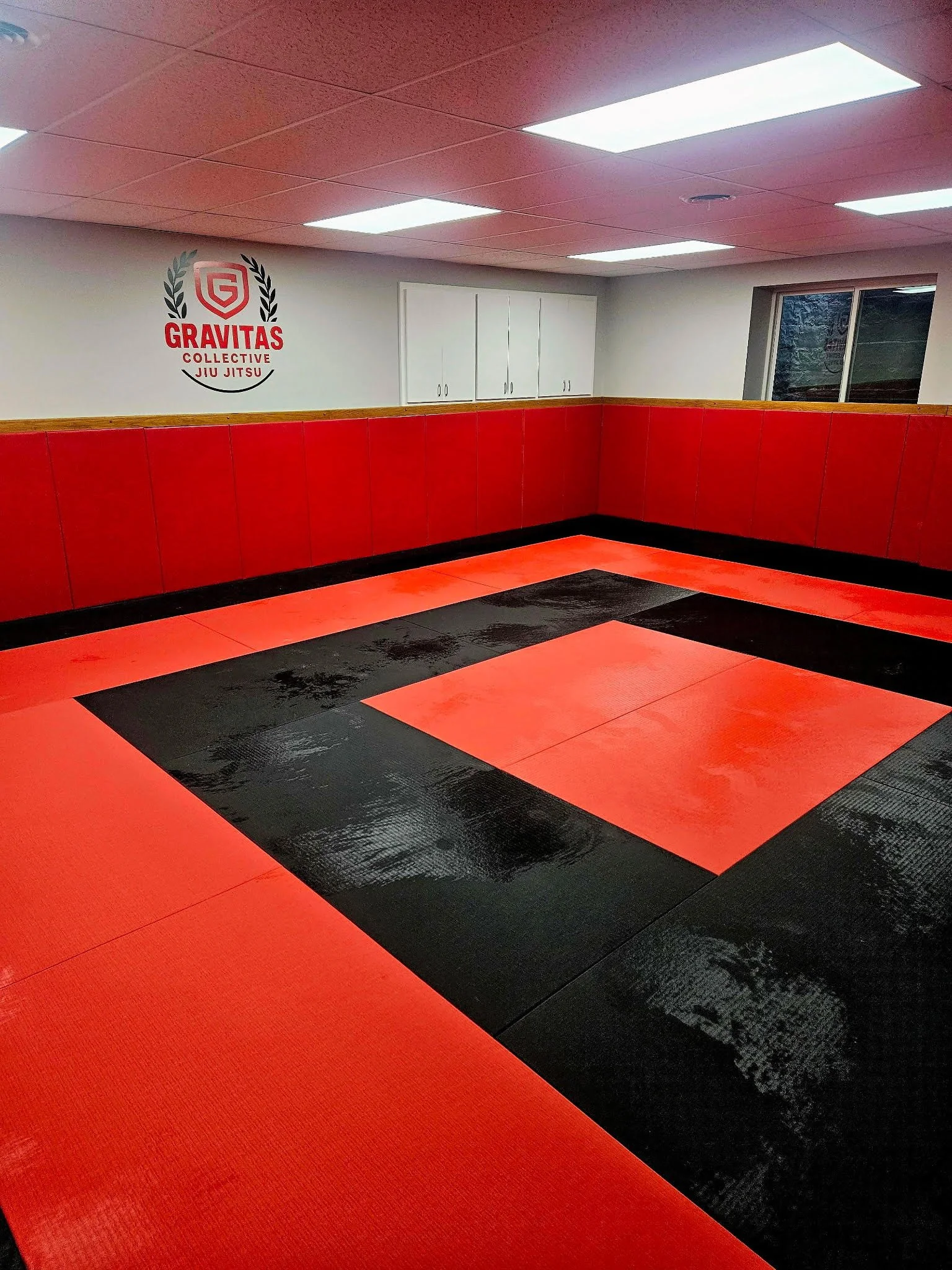 Interior of a martial arts studio with red and black mats on the floor, red padded walls, white cabinetry, and a wall sign for Gravitas Collective Jiu Jitsu. There is a window on the right side.