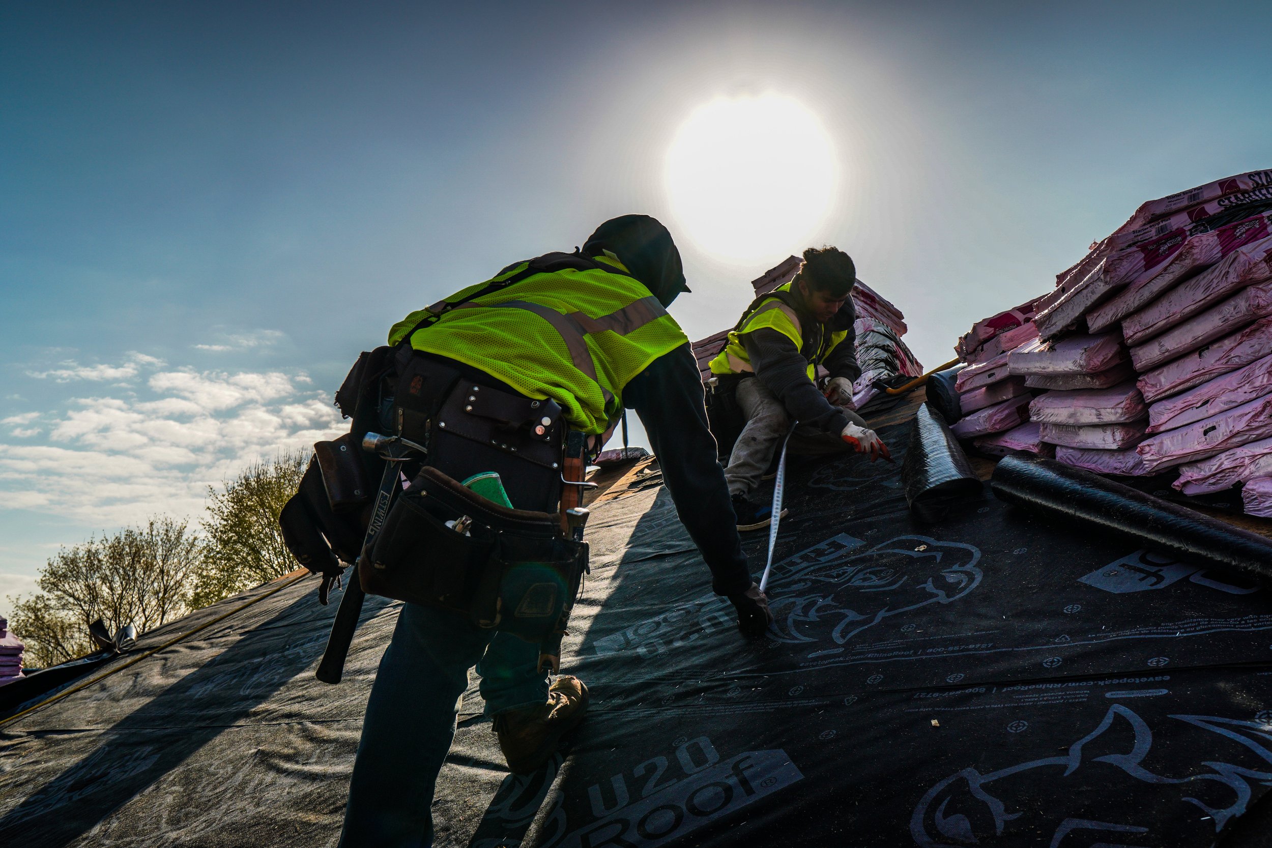 Two construction workers wearing high-visibility safety vests installing roofing underlayment on a roof during daytime with the sun shining.