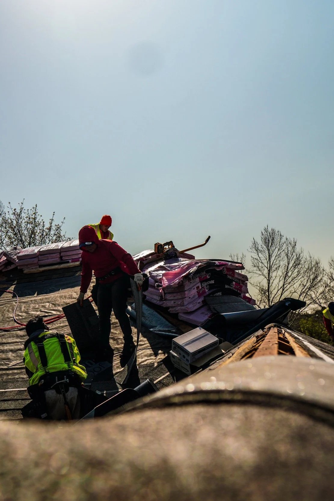 Workers on a roof installing or repairing insulation and equipment, with trees and clear sky in the background.