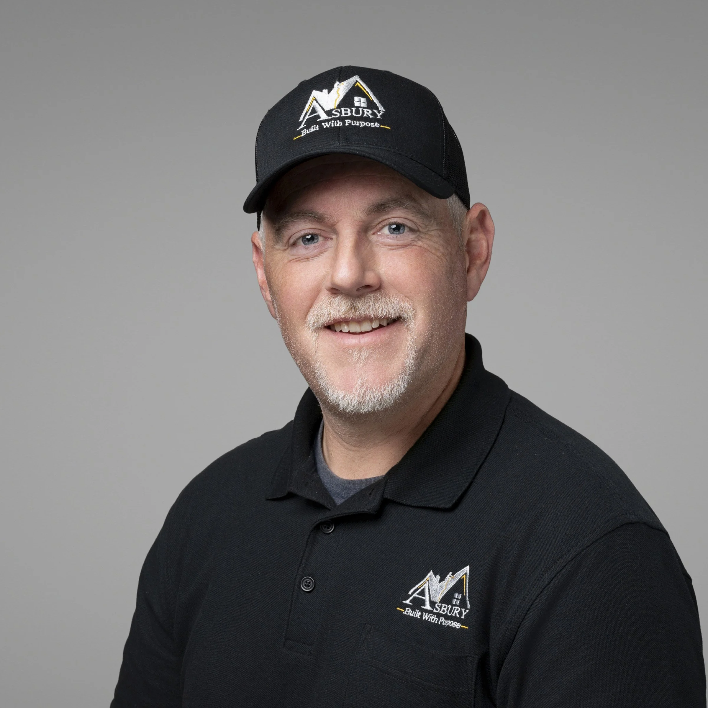 Close-up of a man's face wearing a black cap and black shirt with an 'Asbury Built With Purpose' logo. The background is plain gray.