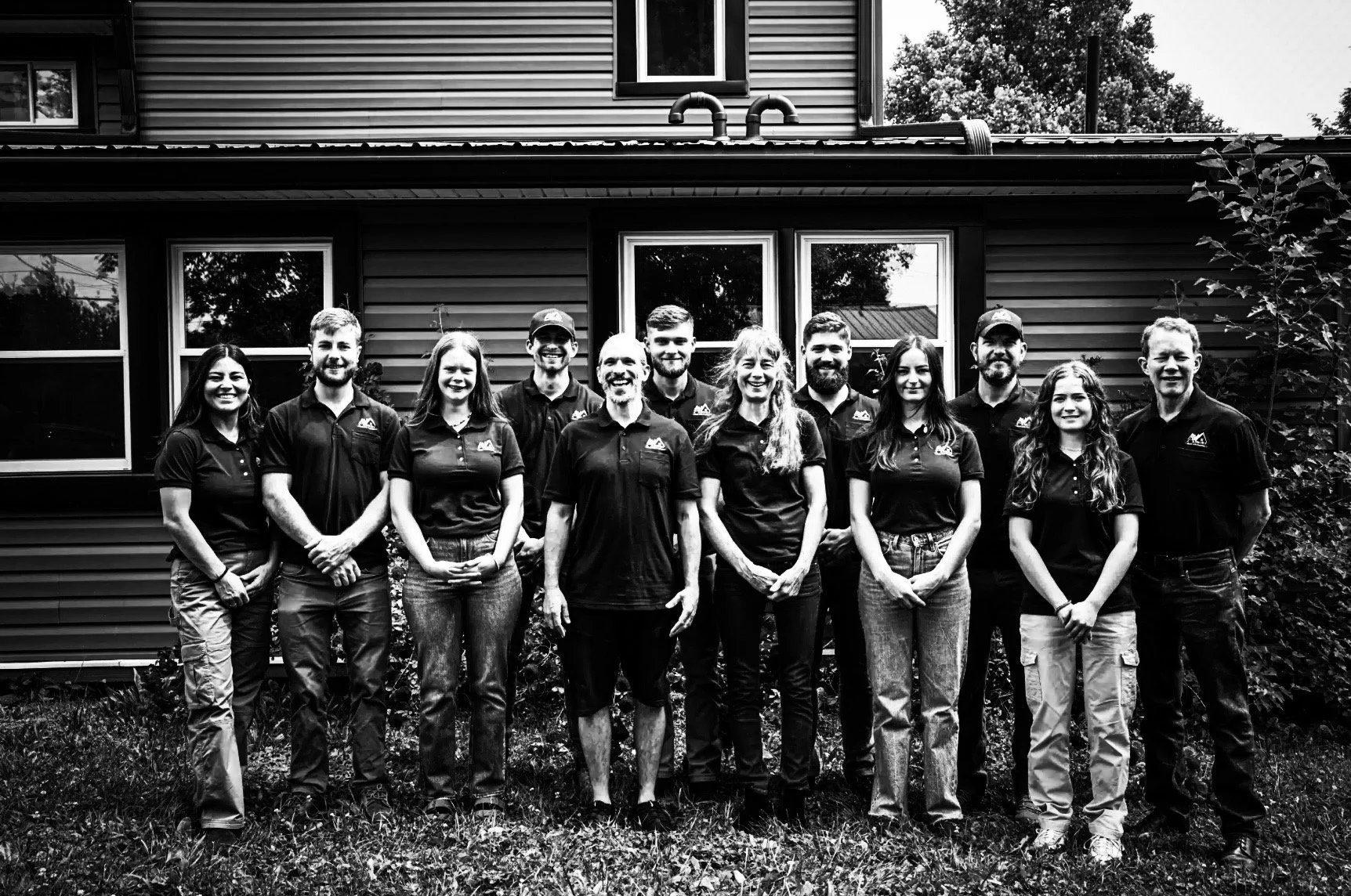 A group of thirteen people standing outdoors in front of a house, smiling at the camera. They are all wearing matching black shirts with a logo, likely colleagues or a team.