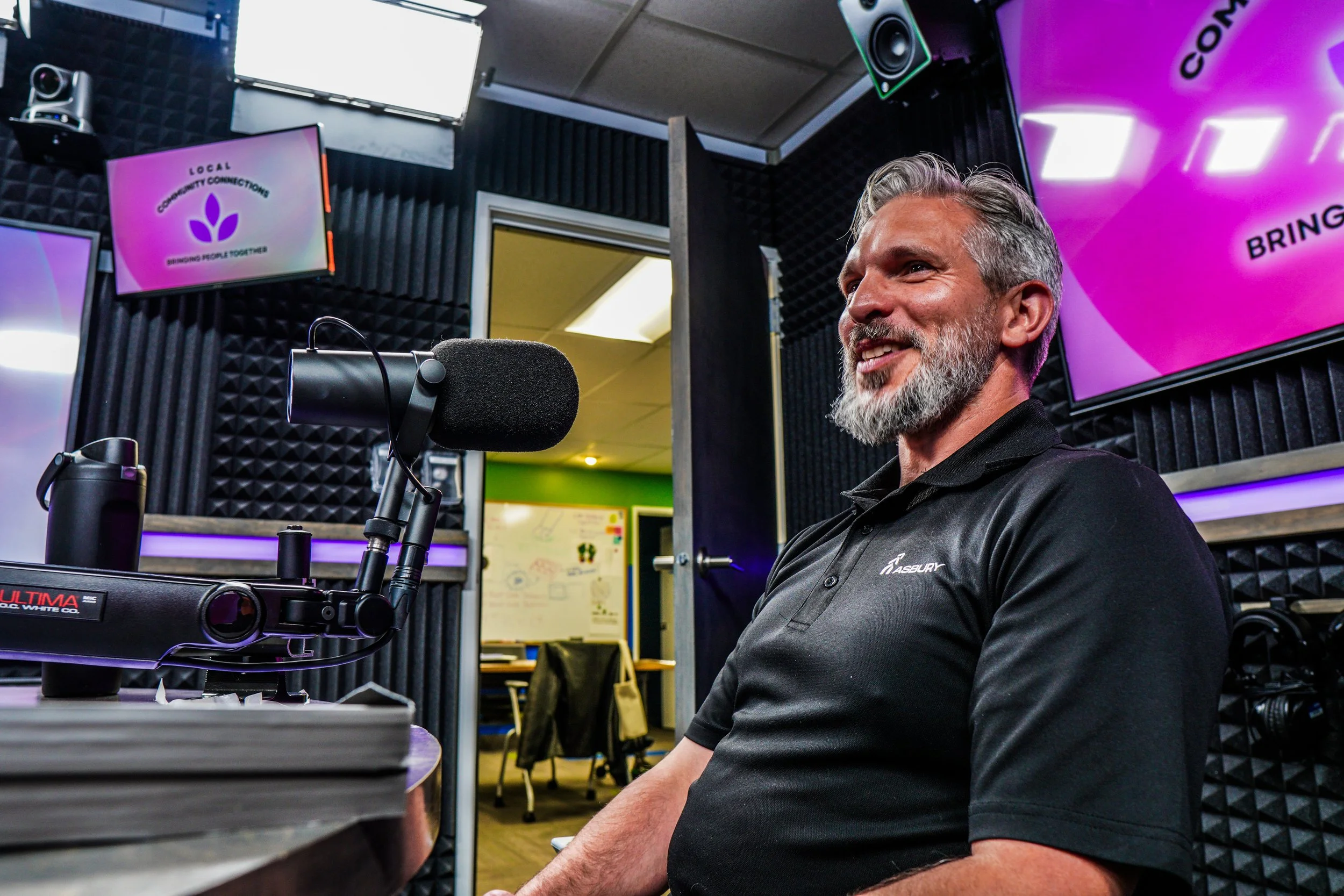 A man with gray hair and beard smiling while sitting in a recording studio with audio equipment and multiple monitors on the walls.
