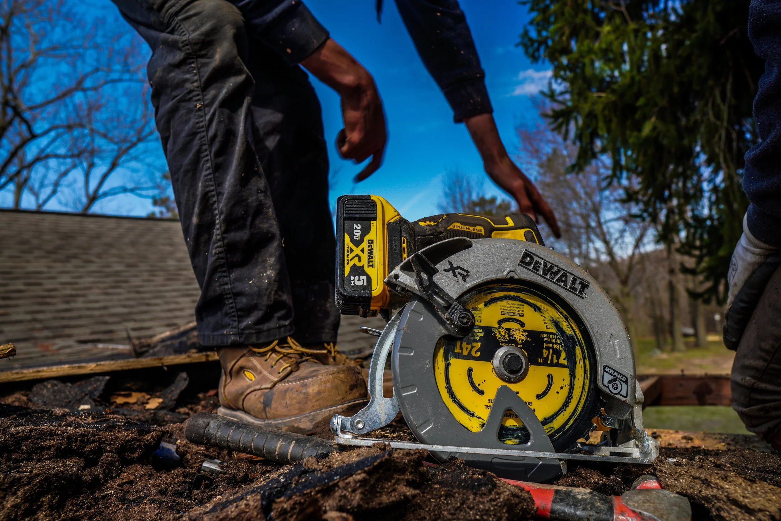 Person using a yellow and silver DEWALT circular saw with a battery pack to cut a piece of wood outdoors, with trees and a blue sky in the background.