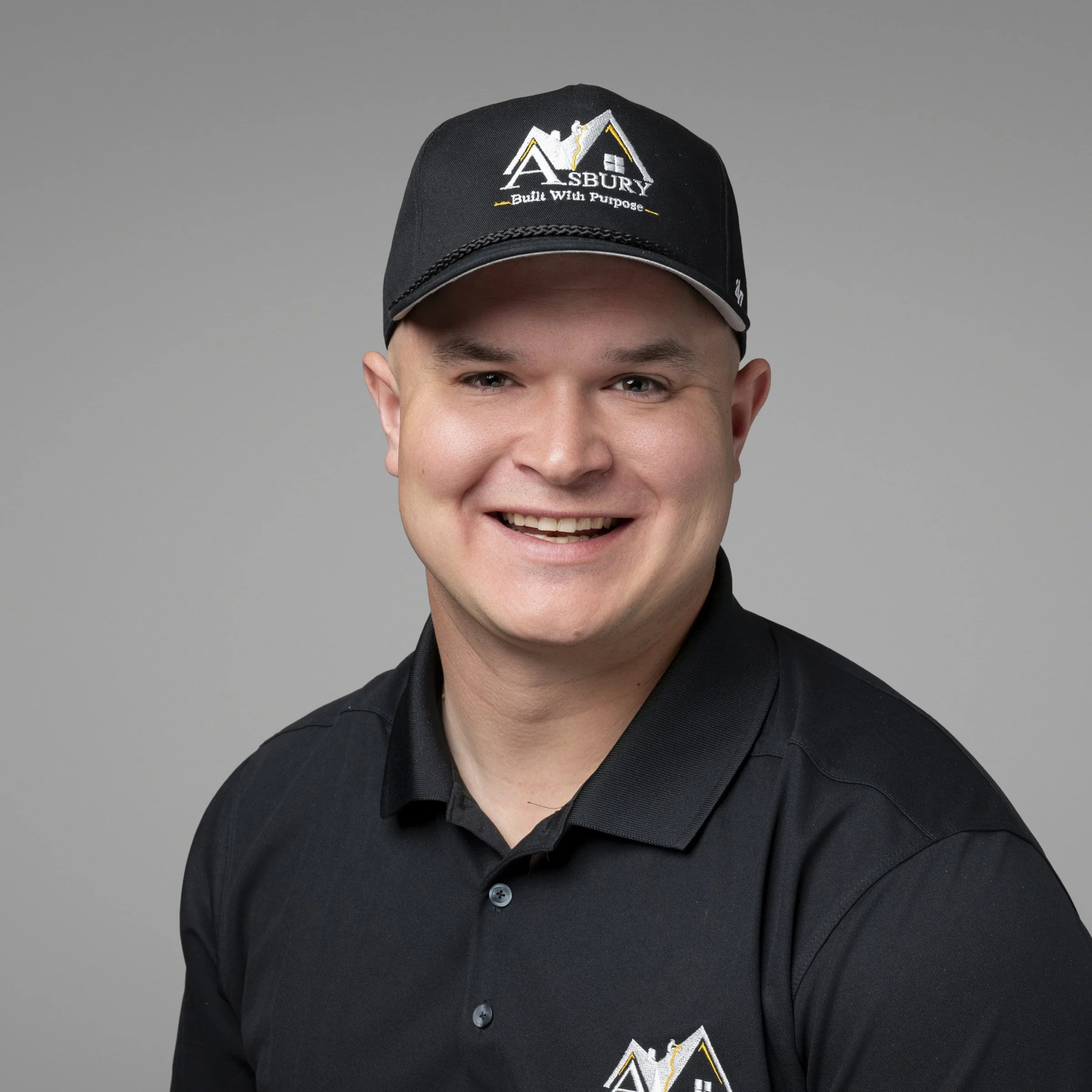 Close-up of a young man smiling wearing a black baseball cap with the logo and text 'Asbury Built With Purpose' embroidered on it.