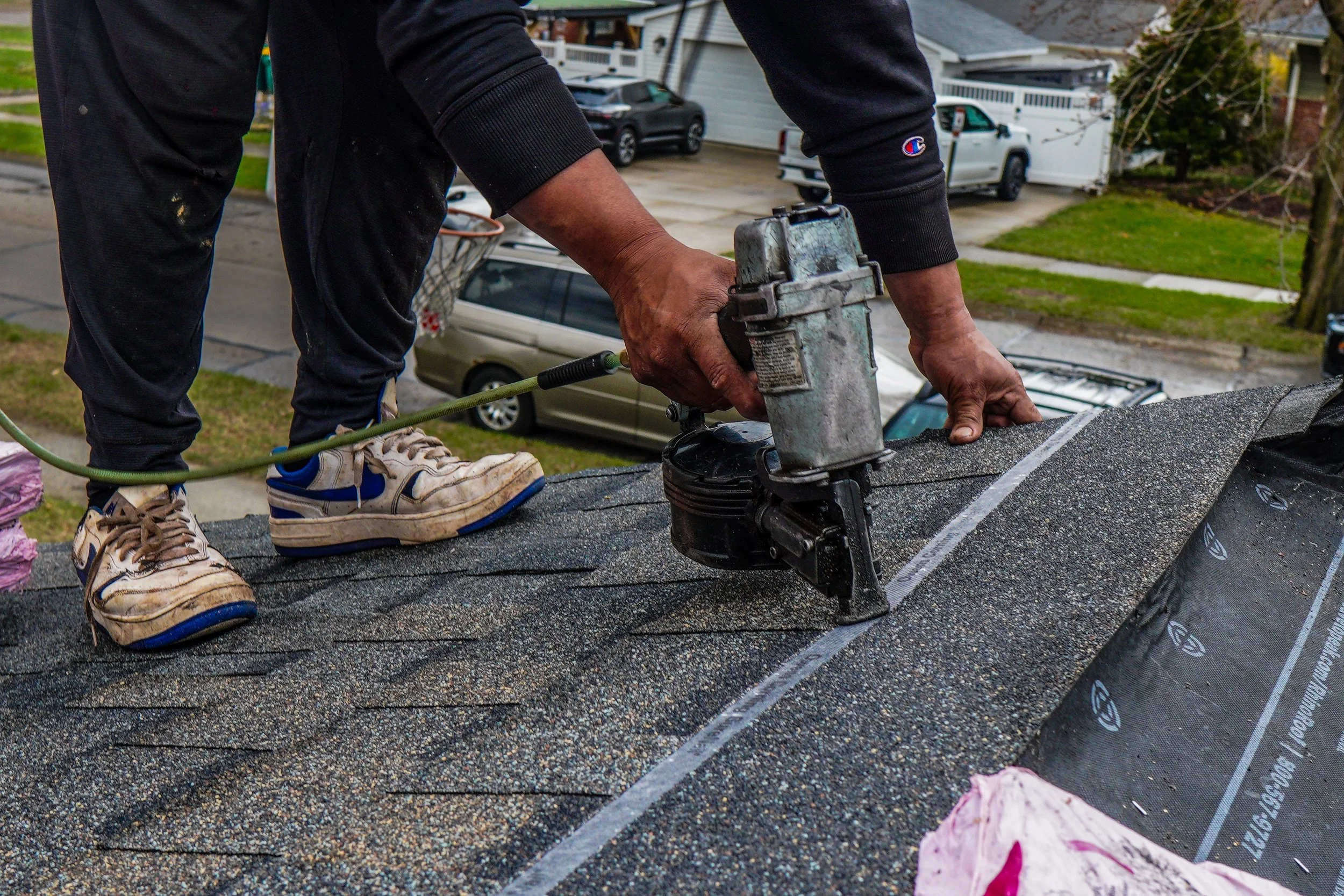 A person working on a roof with a roofing nail gun, wearing sneakers, black pants, and a black hoodie, with trees and parked cars visible in the background.