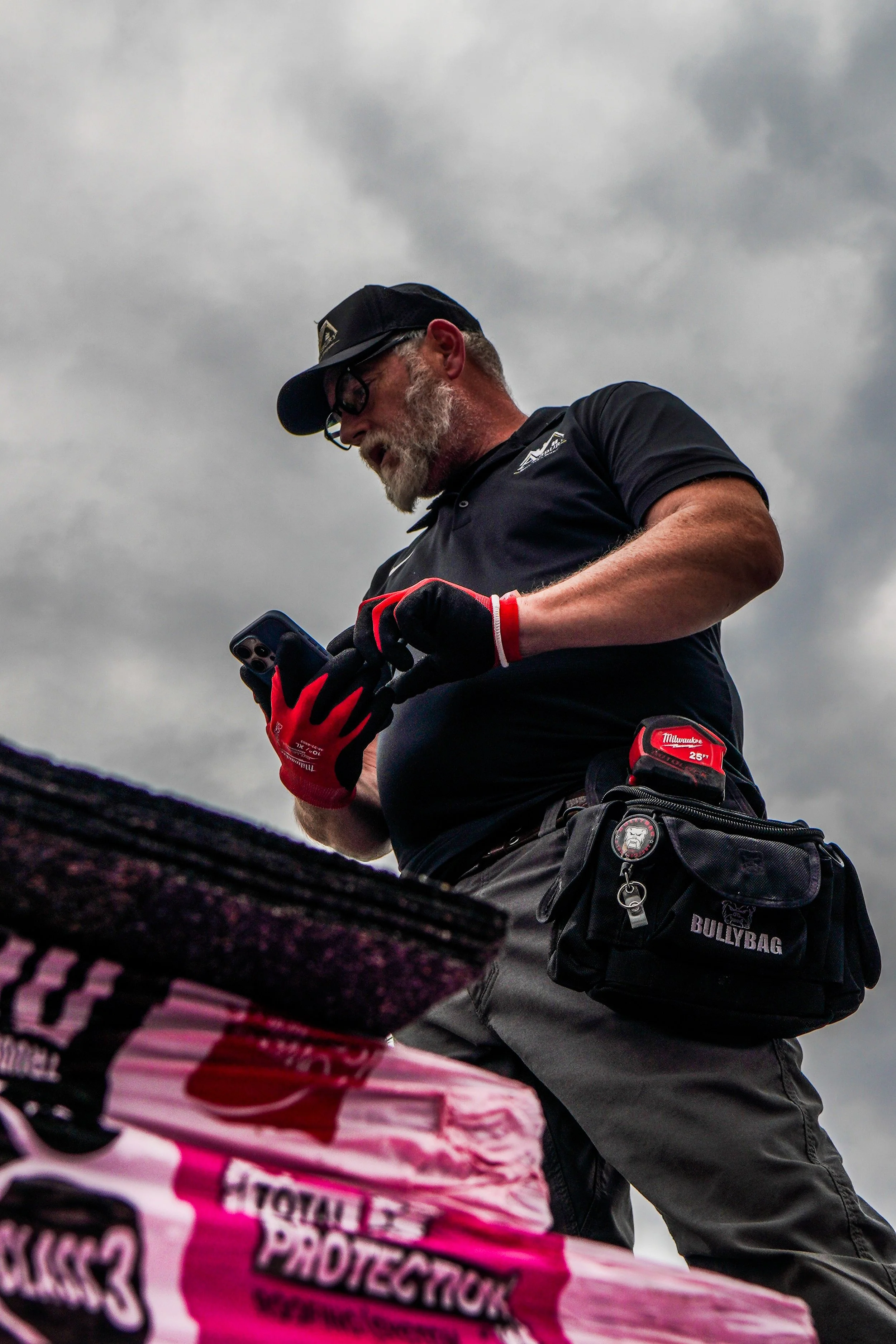 A man wearing glasses, a black cap, and black work gloves is looking at his phone outdoors under a cloudy sky. He is dressed in a dark shirt and has a utility pouch attached to his waist and a tape measure clipped to it.