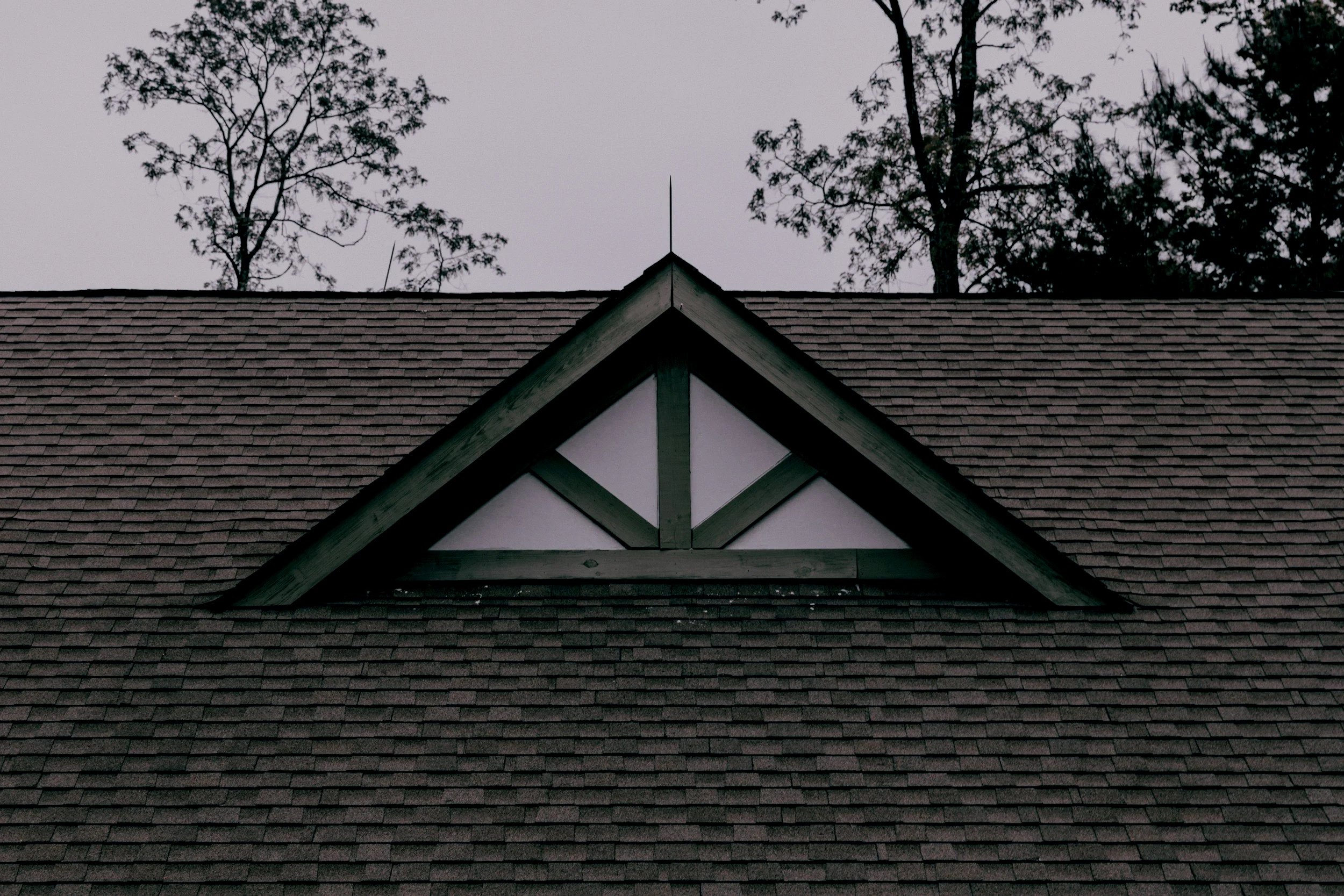 The image shows a close-up view of a house's roof with brown shingles and a triangular gable with decorative trim, against a background of tall trees and an overcast sky.