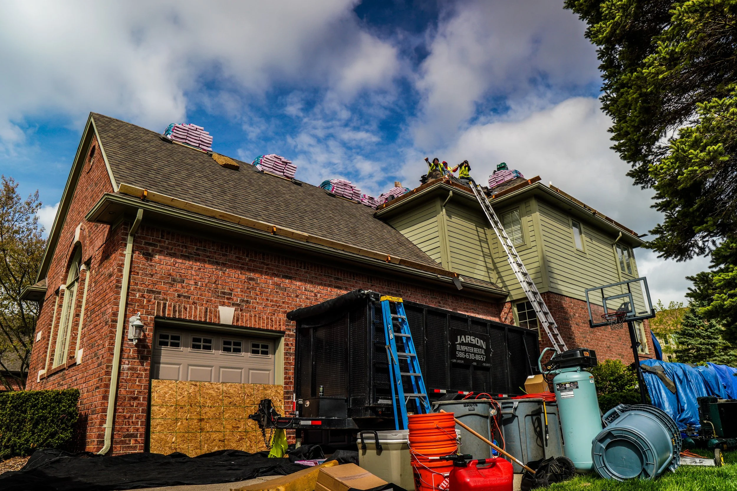 Construction workers working on the roof of a house with stacks of roofing material, ladders, and construction equipment in the yard.