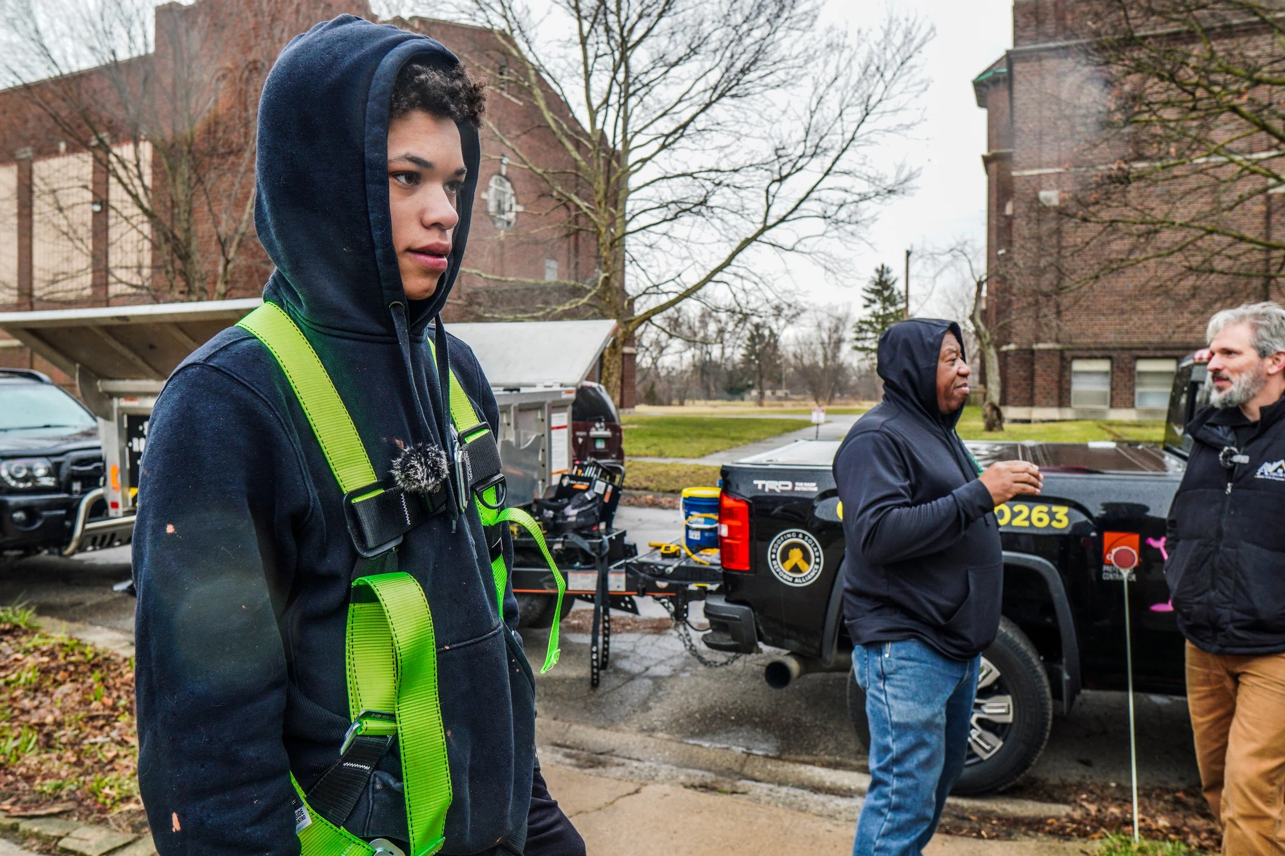 Young man in hoodie and safety harness standing outdoors, with two other individuals near utility trucks and equipment, trees, and brick buildings in the background.