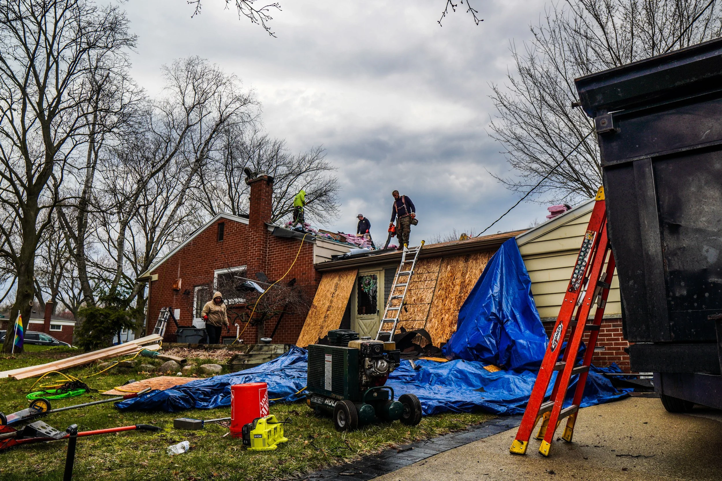 Workers repairing and renovating a house with a damaged roof on a cloudy day. Construction tools and equipment are scattered around the yard, including ladders, power tools, and protective tarps.